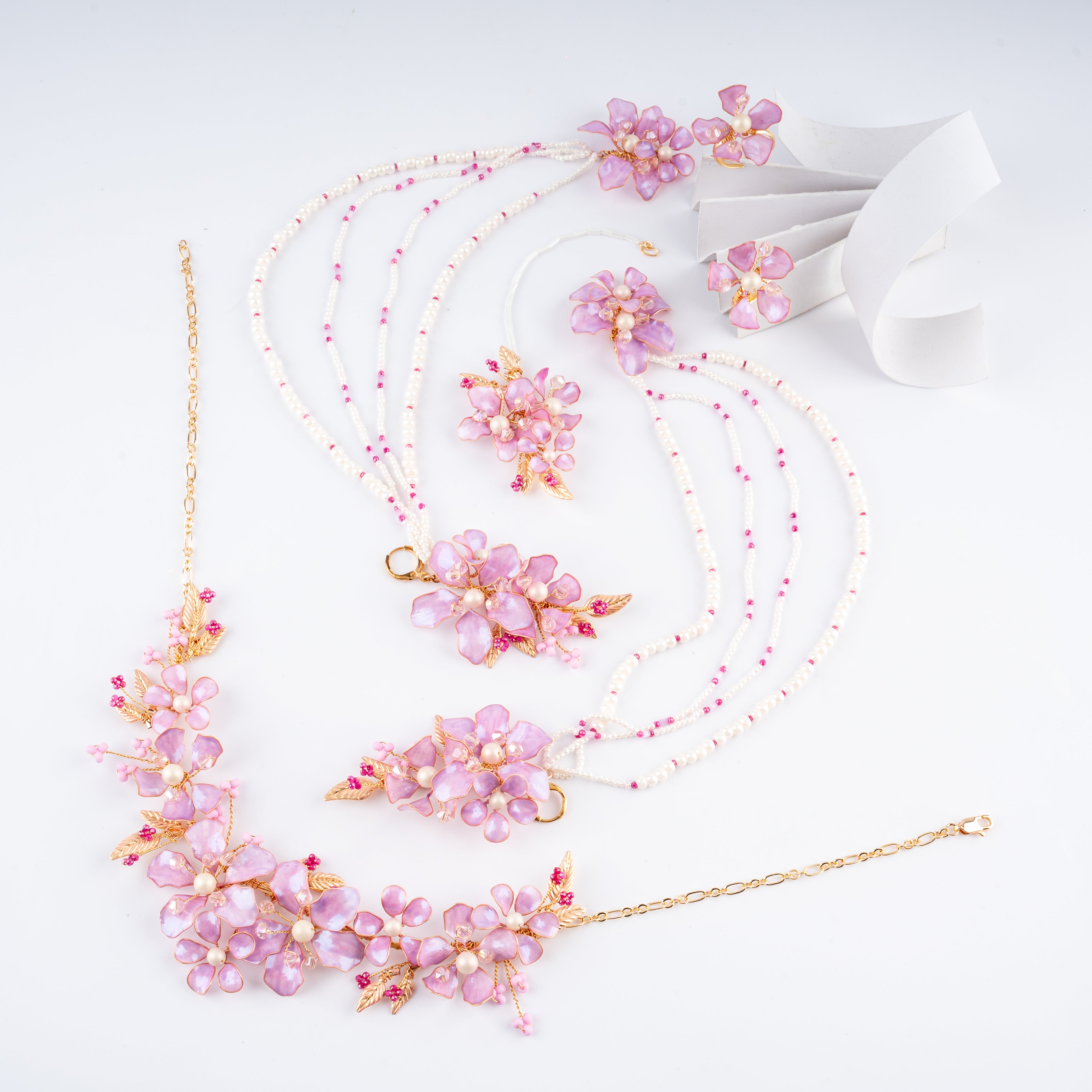 A pink and gold floral jewelry set arranged on a white background, including a beaded necklace, matching earrings, a bracelet, and a white ribbon.