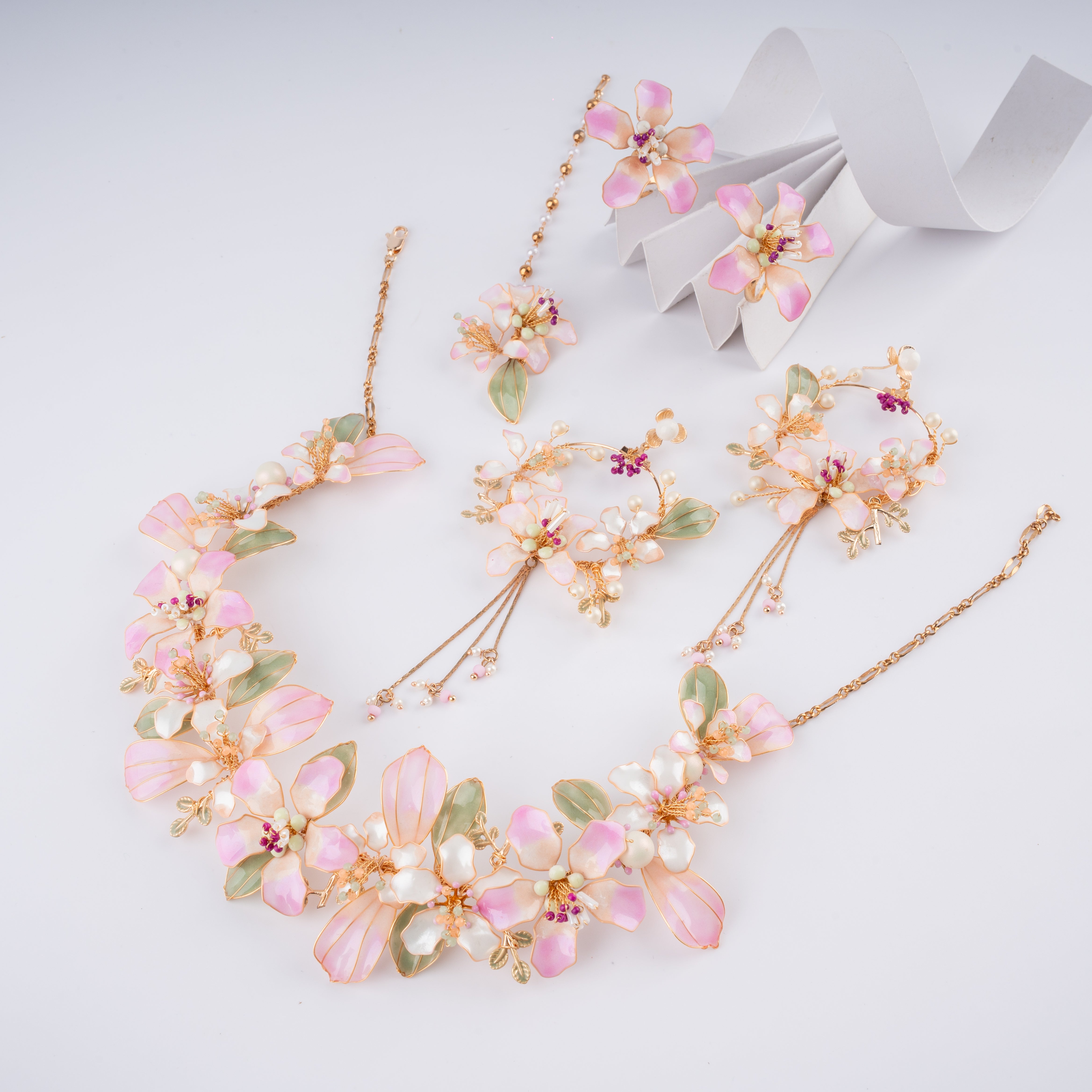 A floral jewelry set on a light gray background, featuring a gold-tone necklace, matching earrings, and a pair of dangling earrings decorated with pink and white flower petals, green leaves, and small purple accents.