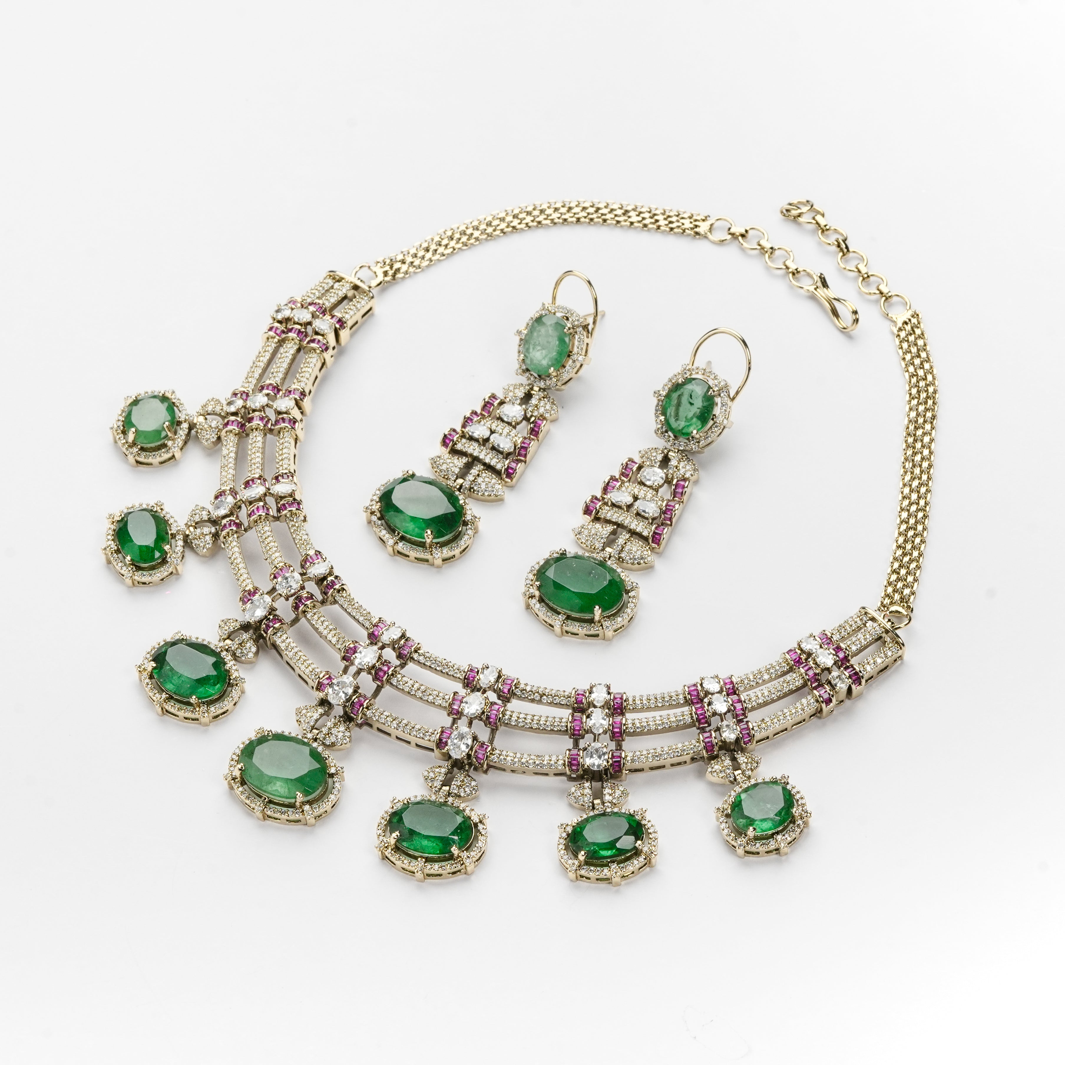 A gold-tone jewelry set featuring a statement necklace and matching drop earrings, embellished with oval green emerald-cut stones and small pink gemstone accents, displayed on a clean white background.