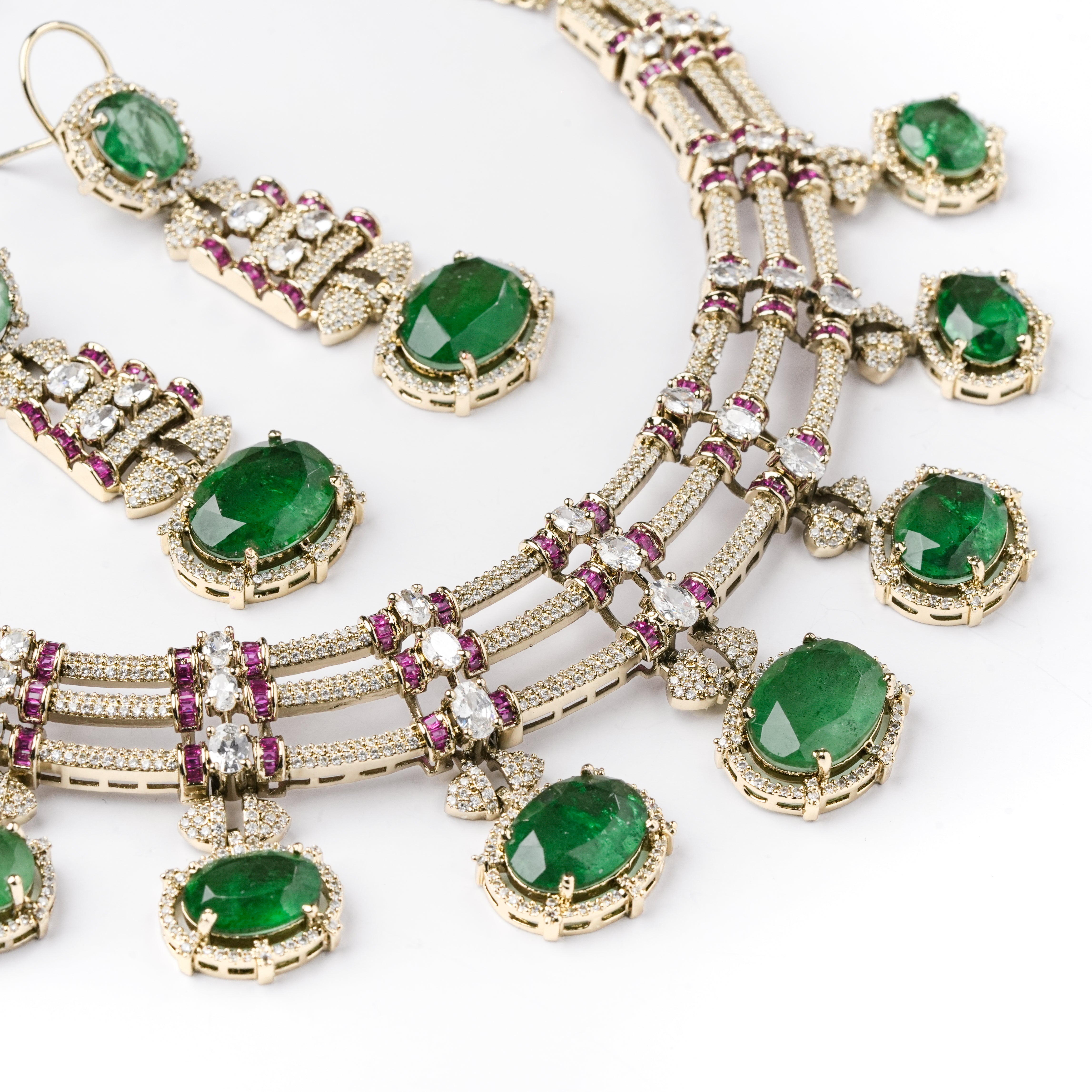 An ornate gold-toned jewelry set featuring a multi-strand necklace and matching earrings, each adorned with oval green emerald-cut gemstones, small clear stones, and pink gemstone accents on a white background.