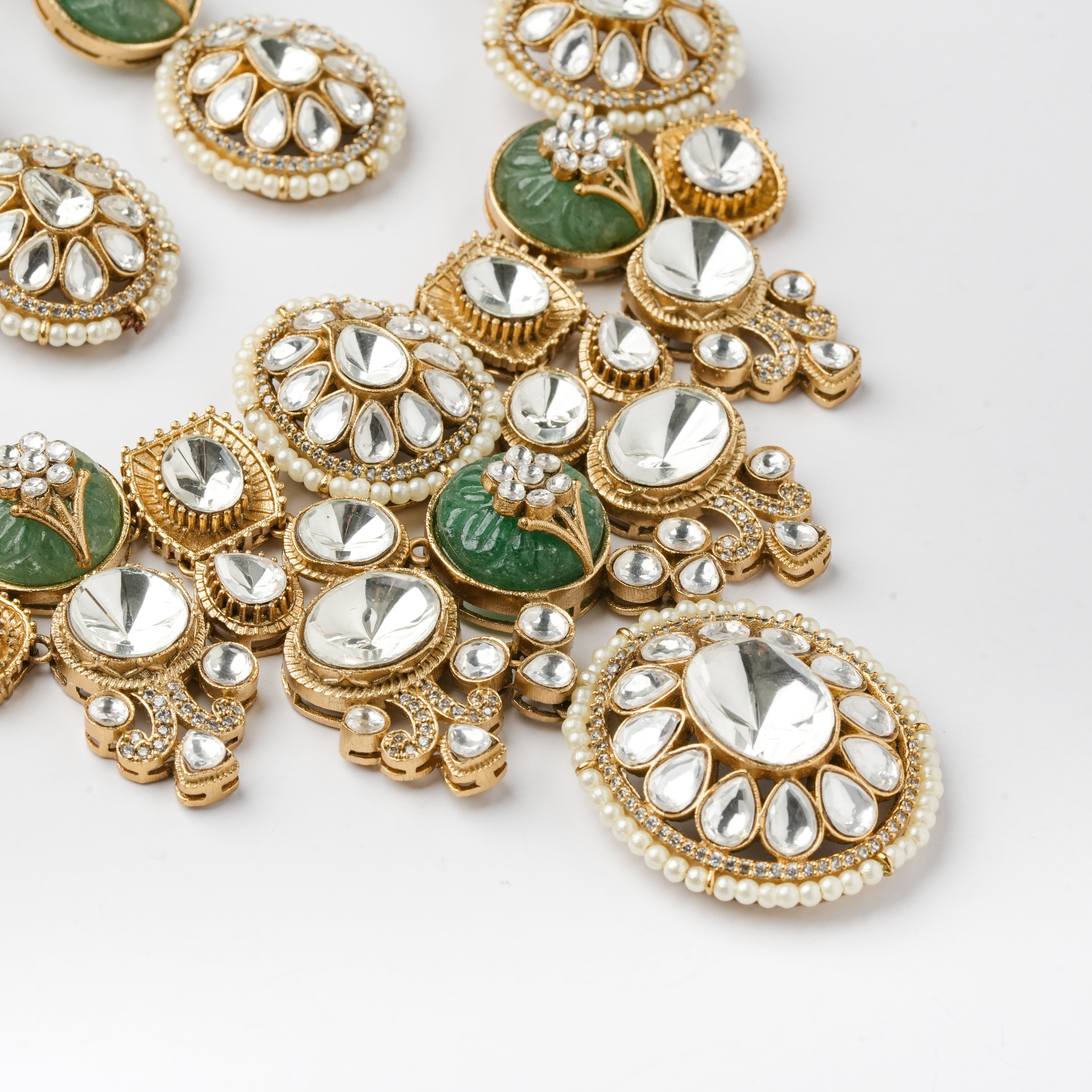 A close-up of ornate gold-toned jewelry pieces with clear teardrop and round stones, small pearl-like beads, and green inlay details on a white background.