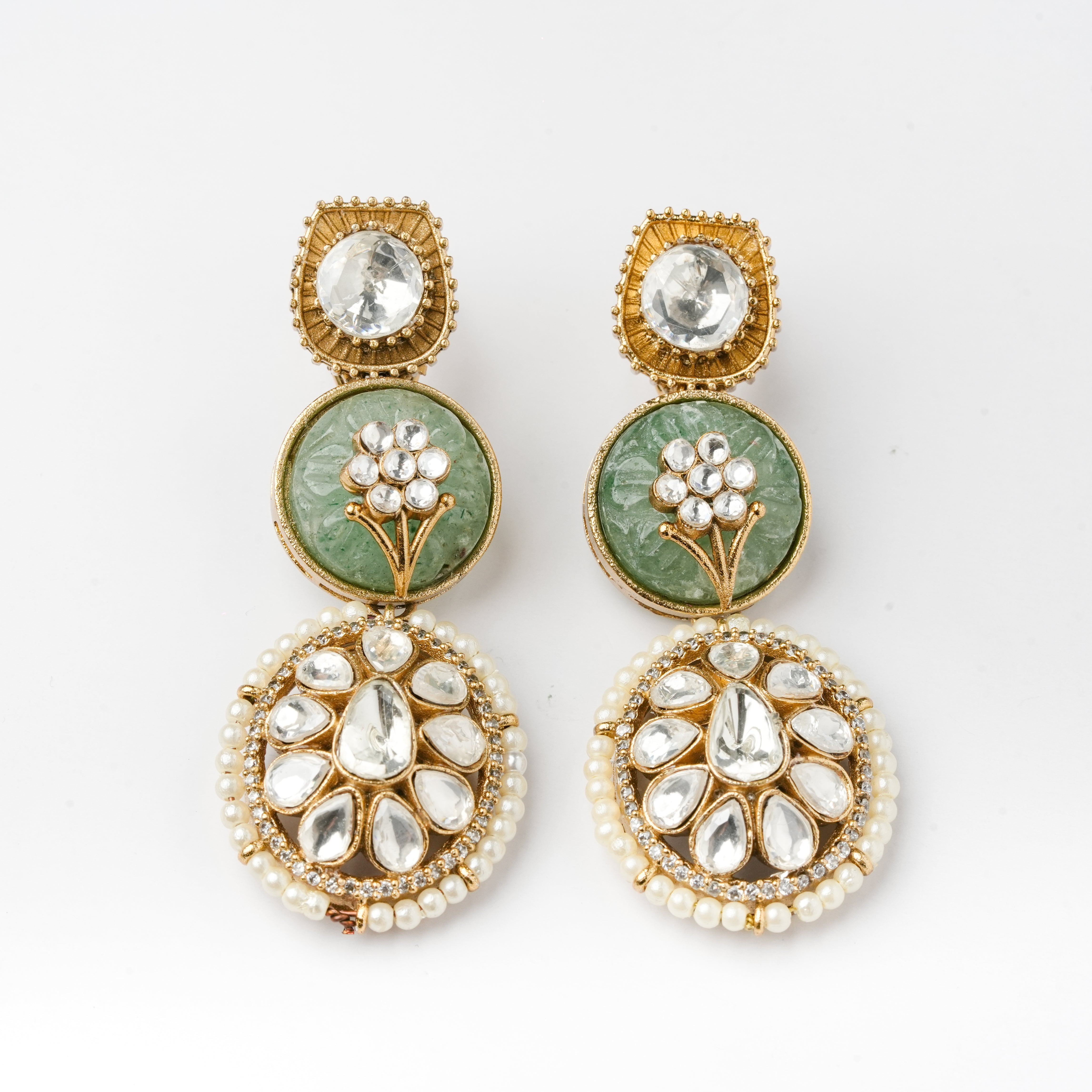 A pair of ornate Indian Kundan earrings featuring green stone inlays, clear crystals, and white faux pearls, set in gold-plated metal on a white background.