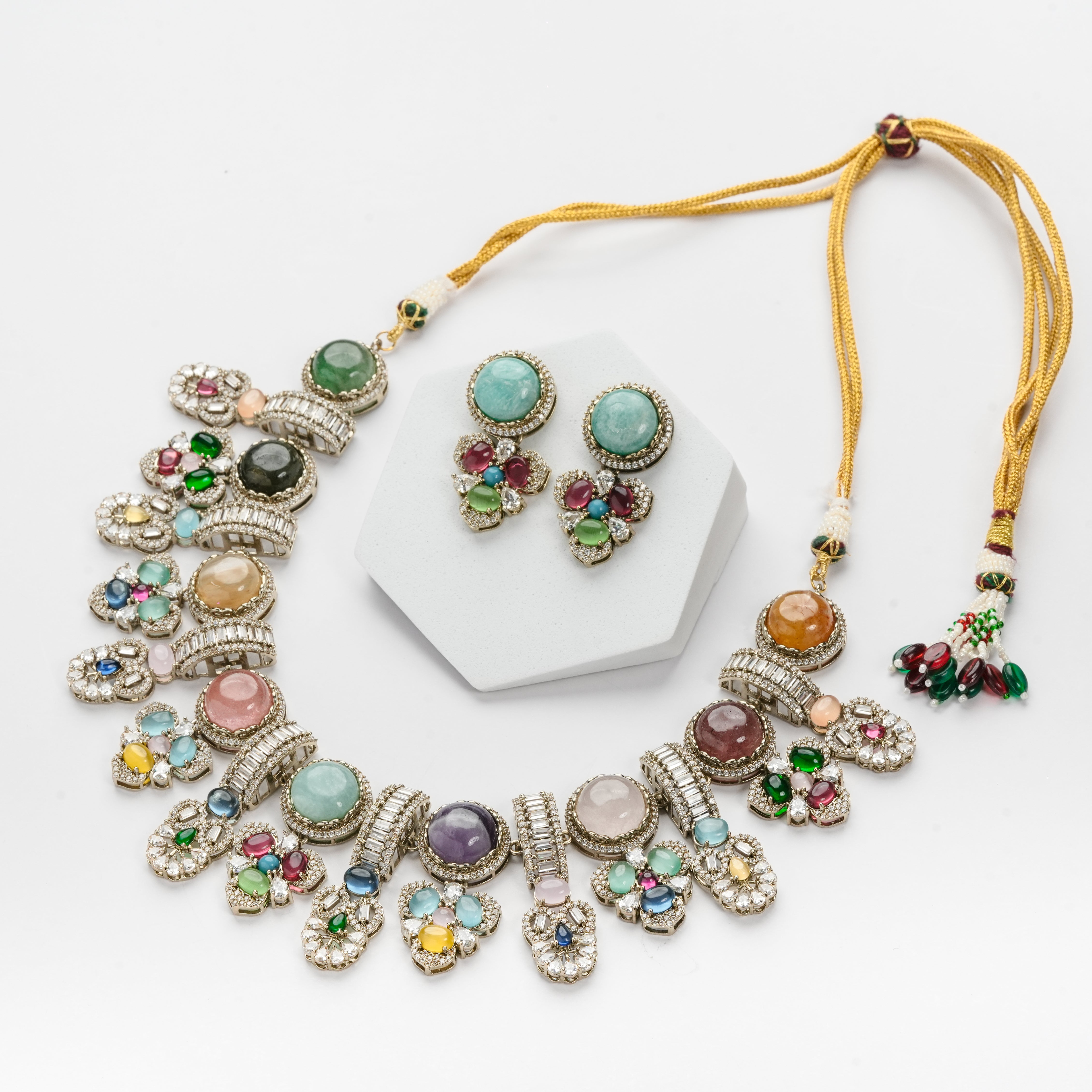 A gold-plated statement necklace and matching stud earrings set with multiple colorful gemstones, displayed on a white background with a small hexagonal stand.