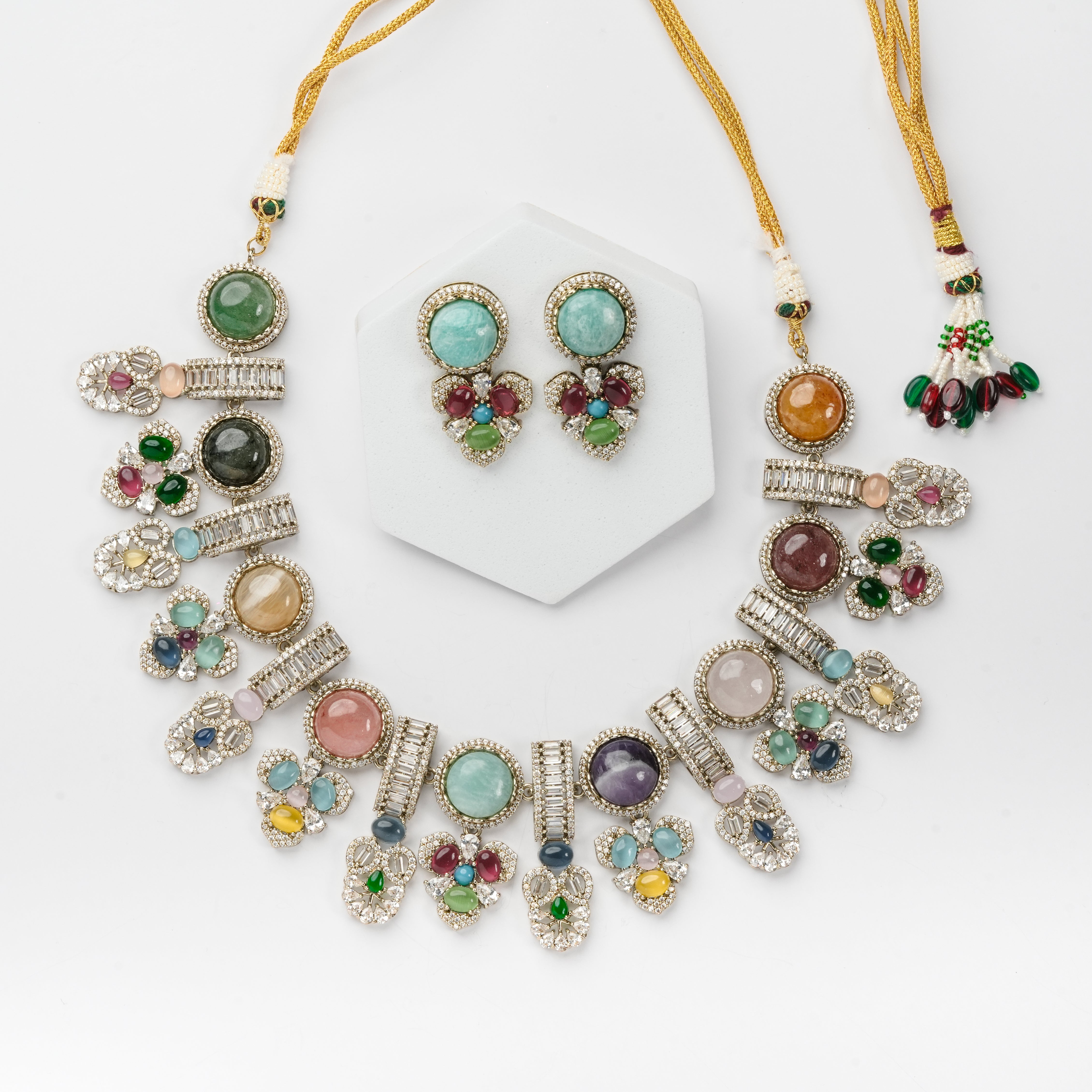 A multicolor gemstone jewelry set featuring a statement necklace and matching stud earrings displayed on a white hexagonal stand against a clean white background.