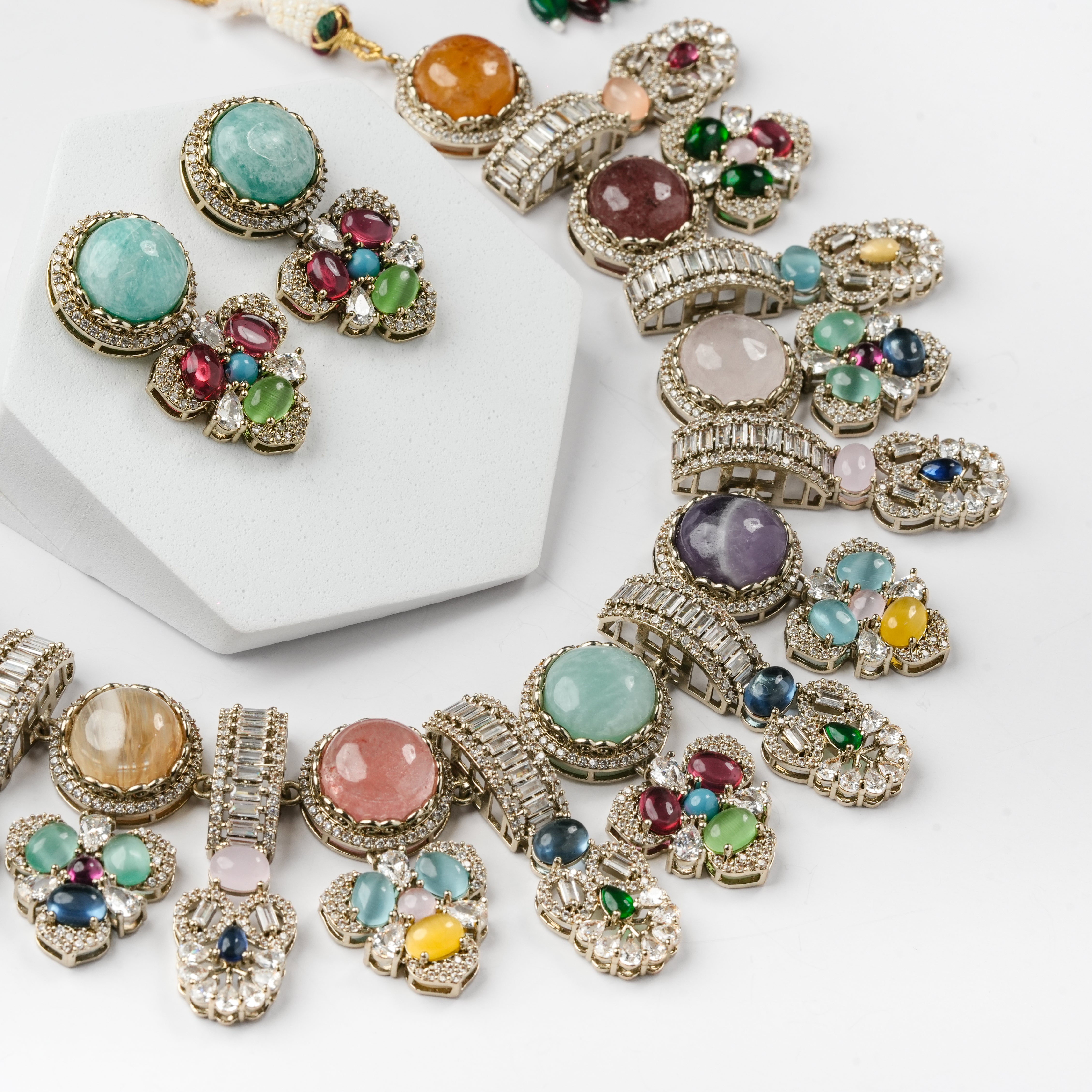 A gold-toned statement jewelry set featuring a chunky statement necklace and matching earrings, embellished with multiple colorful round and oval gemstones and clear rhinestones, displayed on a white background with a white hexagonal stand.