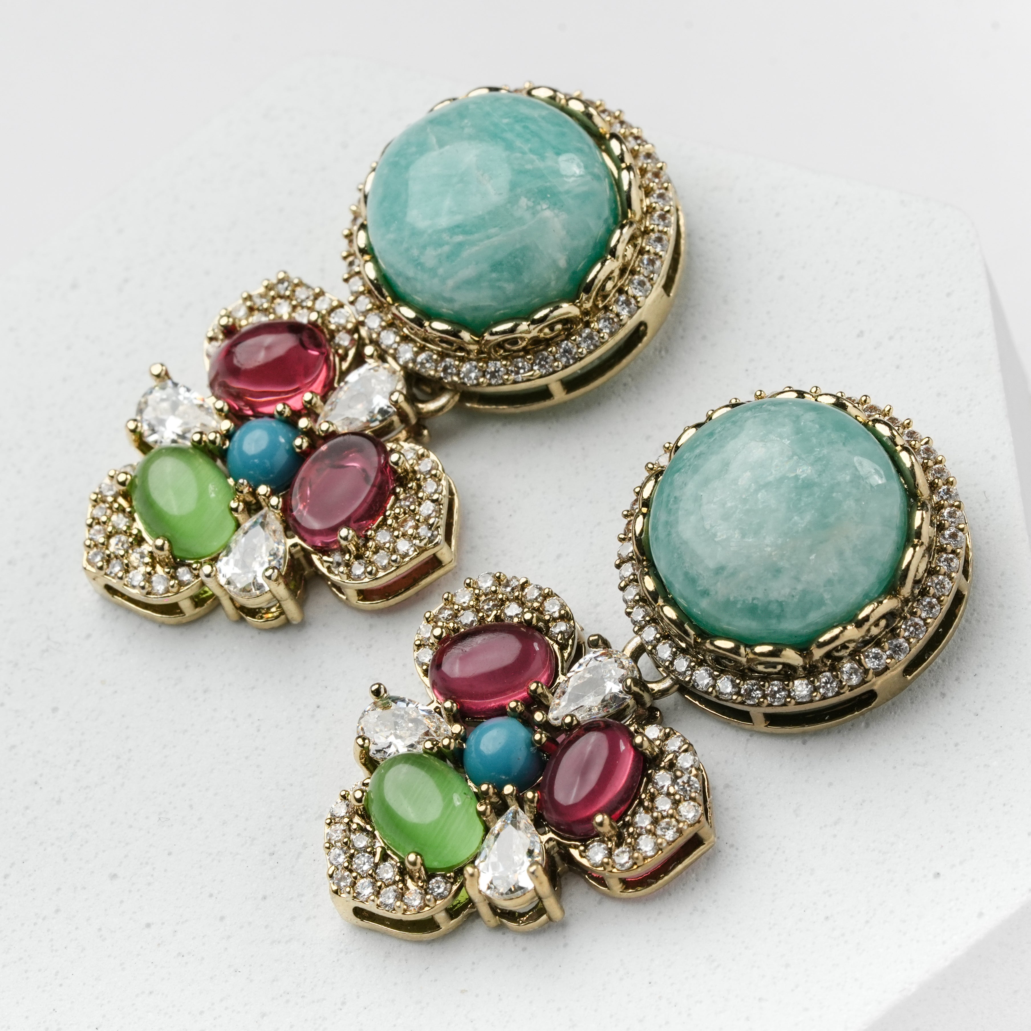 A pair of gold-tone statement earrings featuring large oval light green stones, smaller oval green and pink stones, a small blue stone, and clear crystal accents, displayed on a light gray surface.