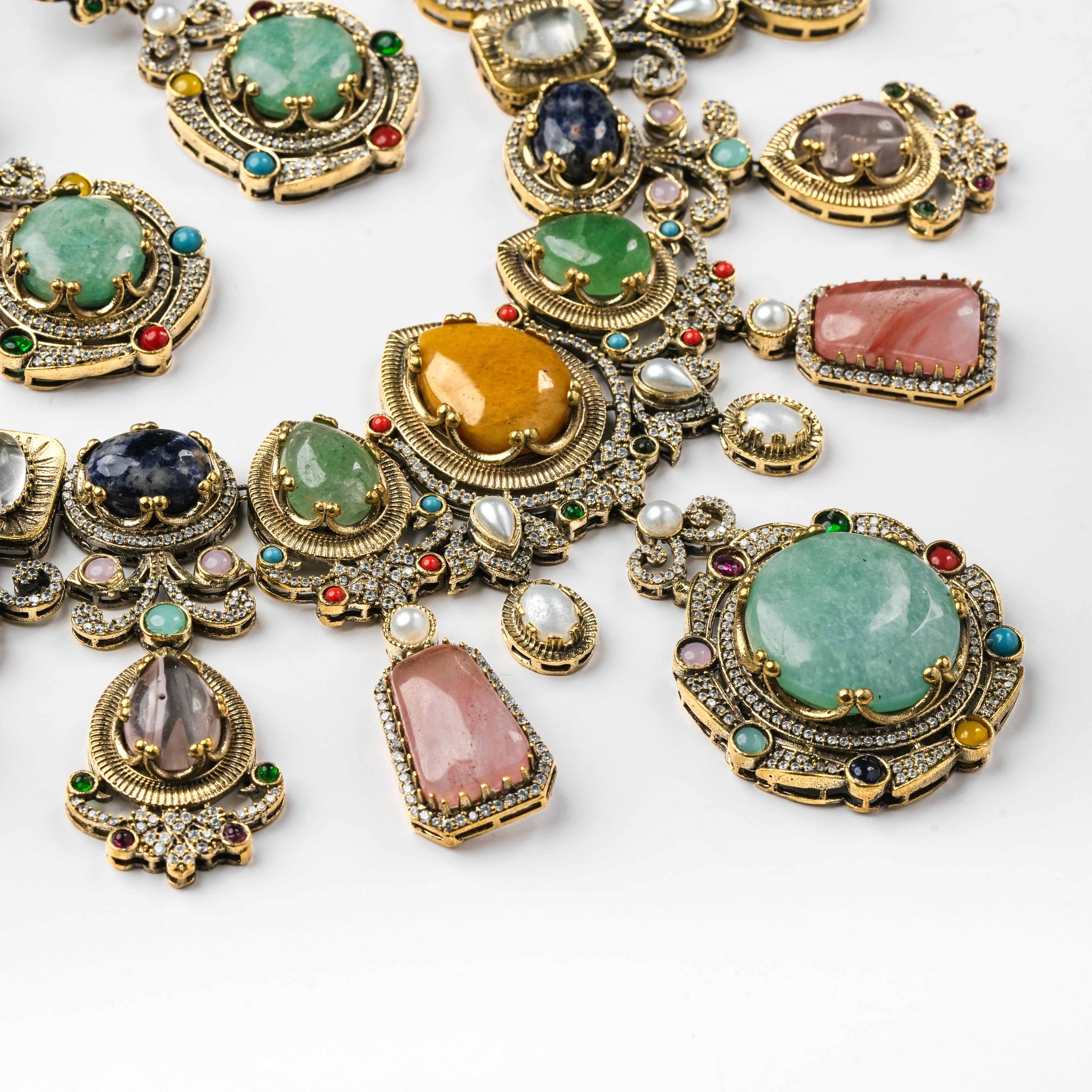 A close-up flat lay of ornate gold-tone jewelry pieces with large multicolored gemstones, including green, pink, orange, blue, and gray stones, set against a plain white background.