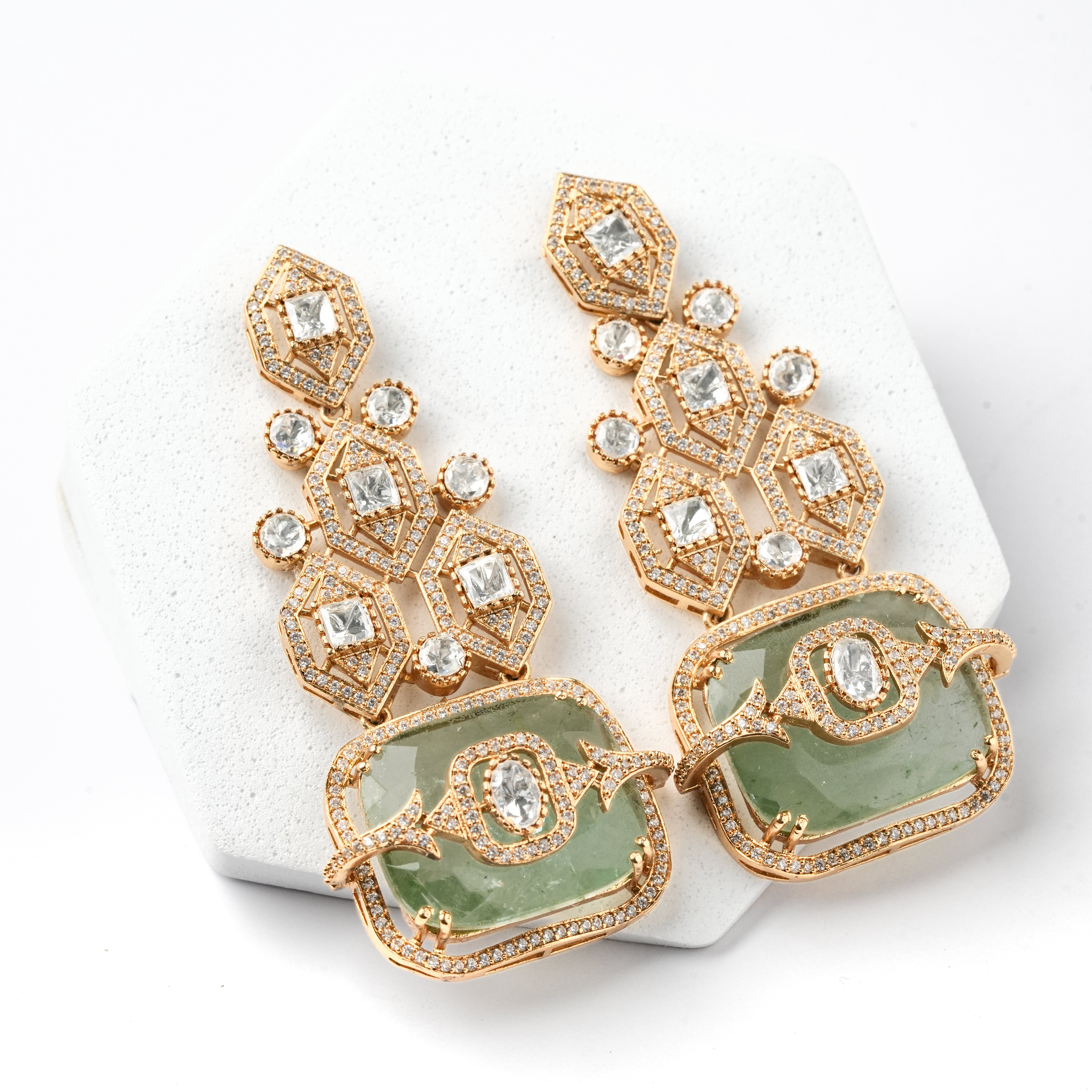 A pair of gold-tone statement earrings with green stone inlays and clear rhinestone accents, displayed on a white textured stand against a white background.