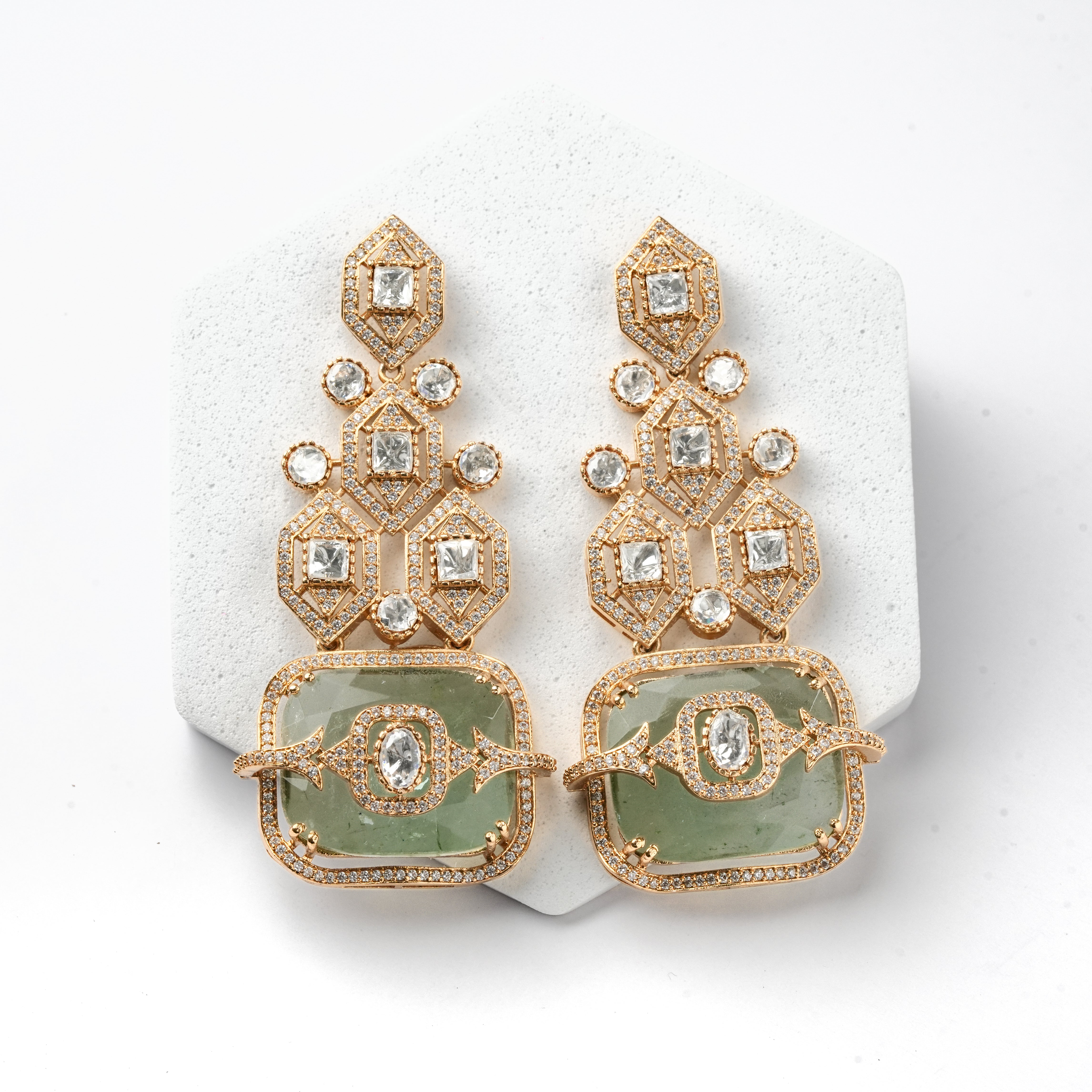 A pair of gold-toned statement earrings with clear gemstones and light green rectangular stone sections, displayed on a white textured hexagonal stand against a white background.