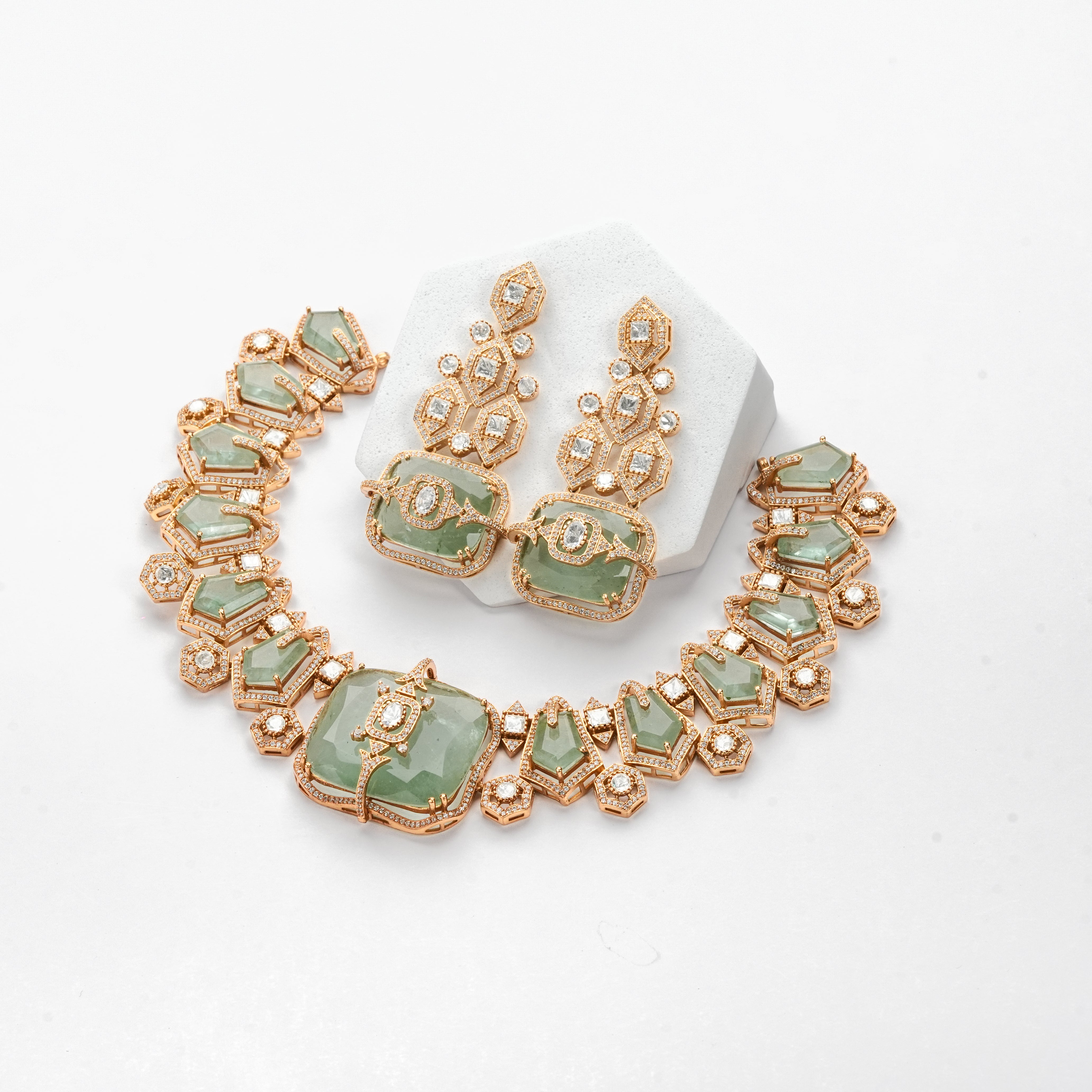 A gold-plated jewelry set featuring a necklace and matching earrings with light green faceted stones and small clear rhinestones, displayed on a white background with a white hexagonal stand.