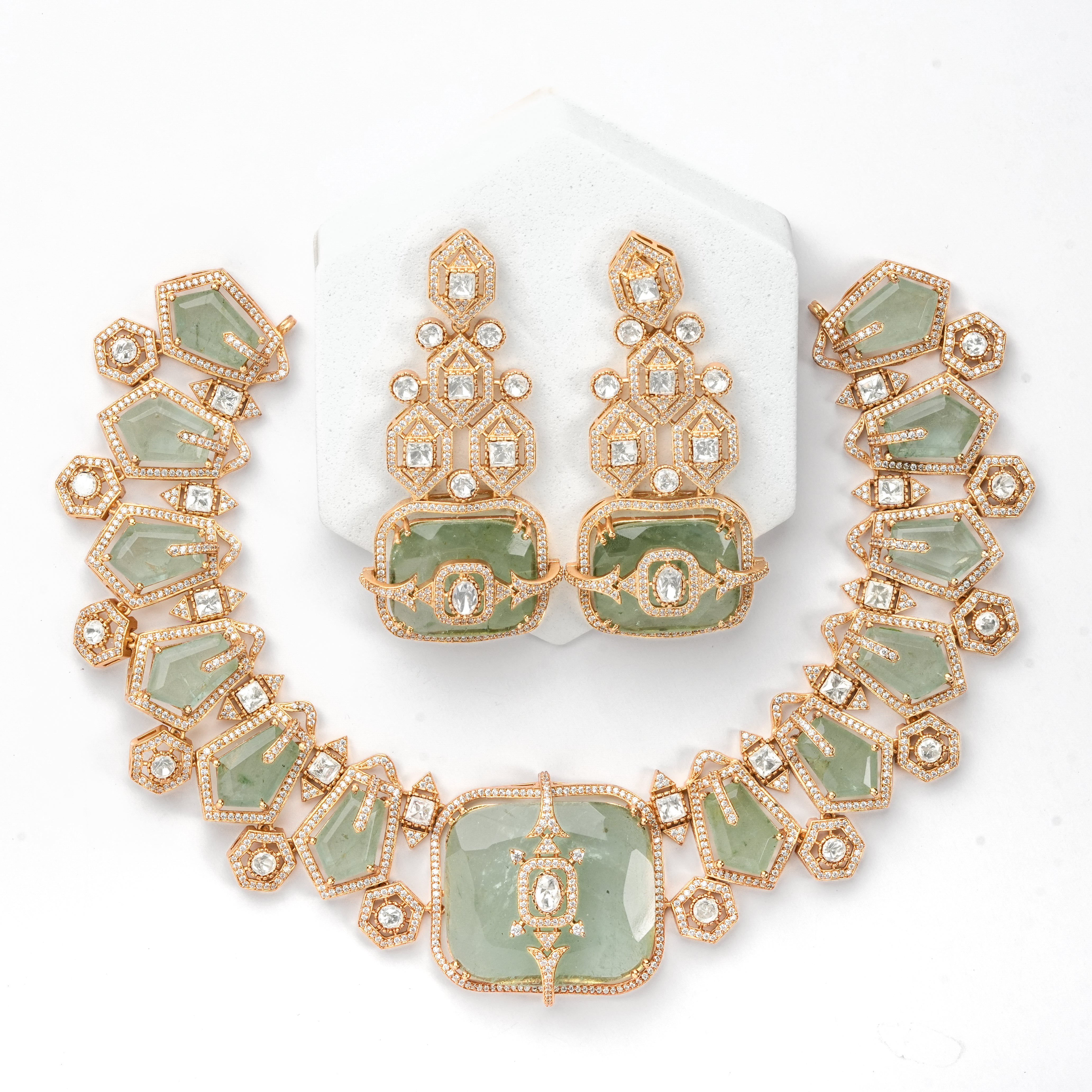 A gold-plated jewelry set featuring a necklace and matching earrings with light green stone inlays and small clear rhinestones, displayed on a white background with a white hexagonal stand.