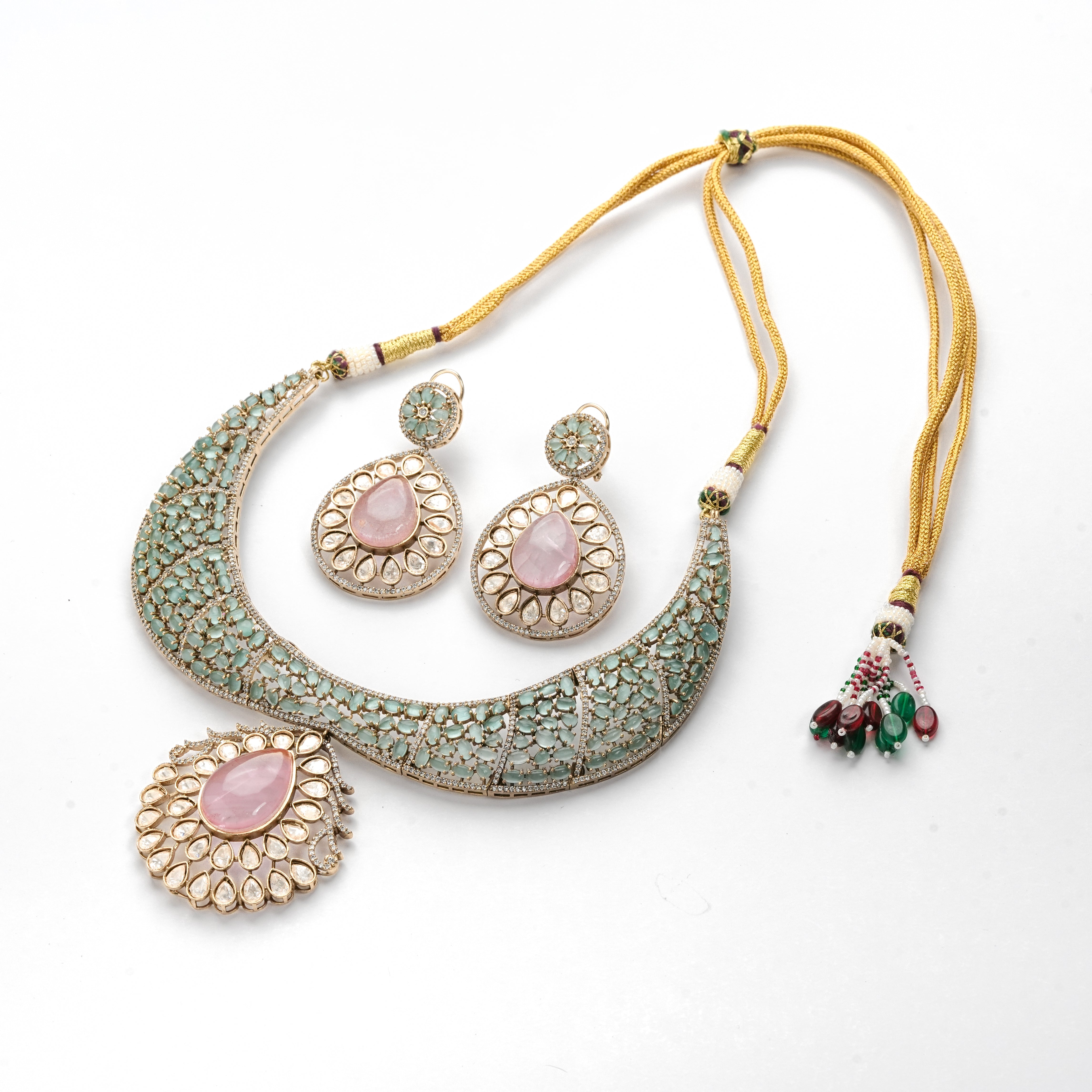 Gold-toned traditional Indian jewelry set featuring a crescent-shaped necklace, a matching pendant, and a pair of teardrop earrings. The pieces are embellished with light green stones and pink stone centers, with a gold cord and tassel details.