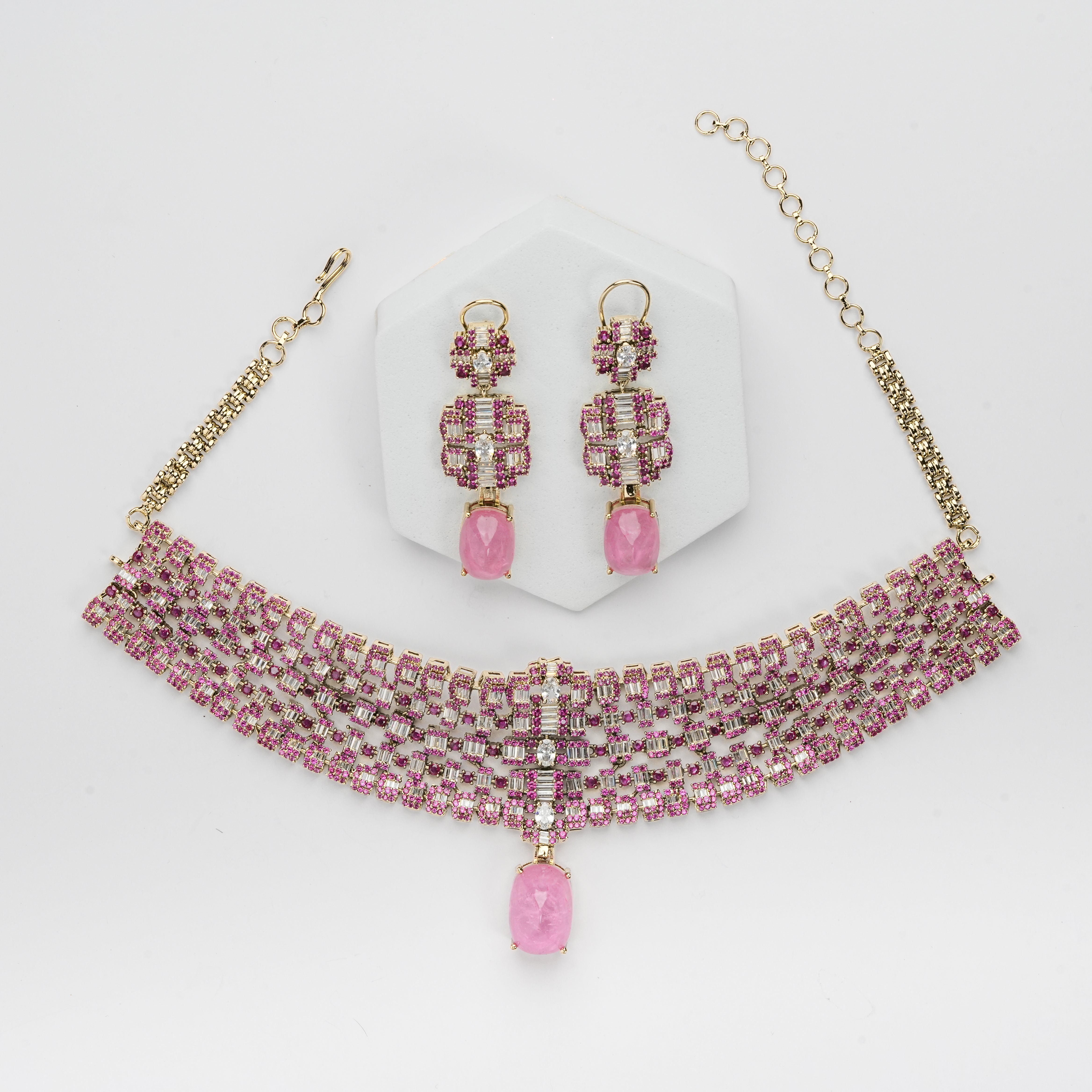 A pink and gold jewelry set featuring a multi-strand beaded choker necklace with a dangling pink teardrop charm, matching drop earrings, and a white hexagonal display stand on a white background.