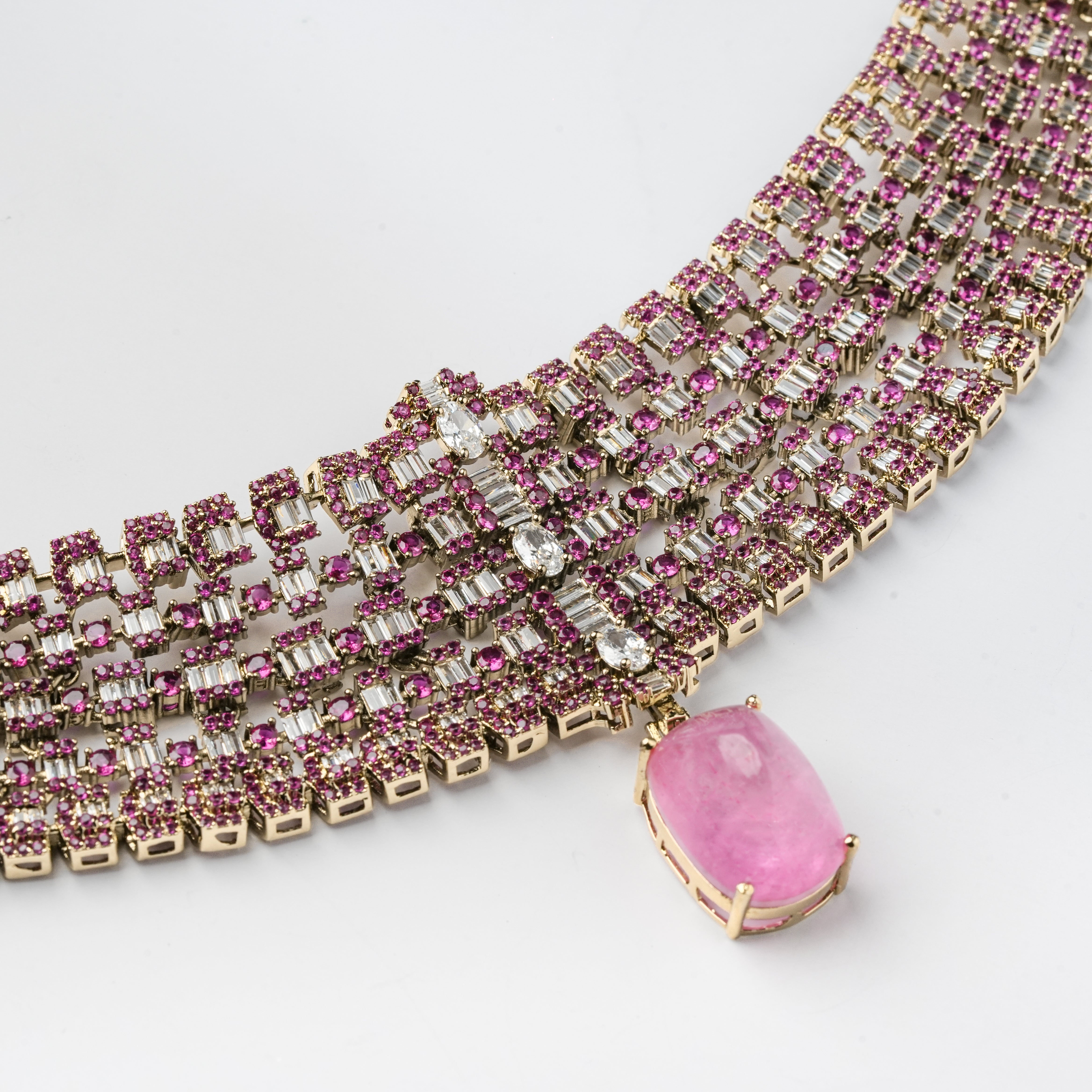 A close-up of a gold-toned multi-row necklace or bracelet embellished with small pink gemstones and clear baguette stones, featuring a large oval pink pendant hanging from one end, set against a plain light background.