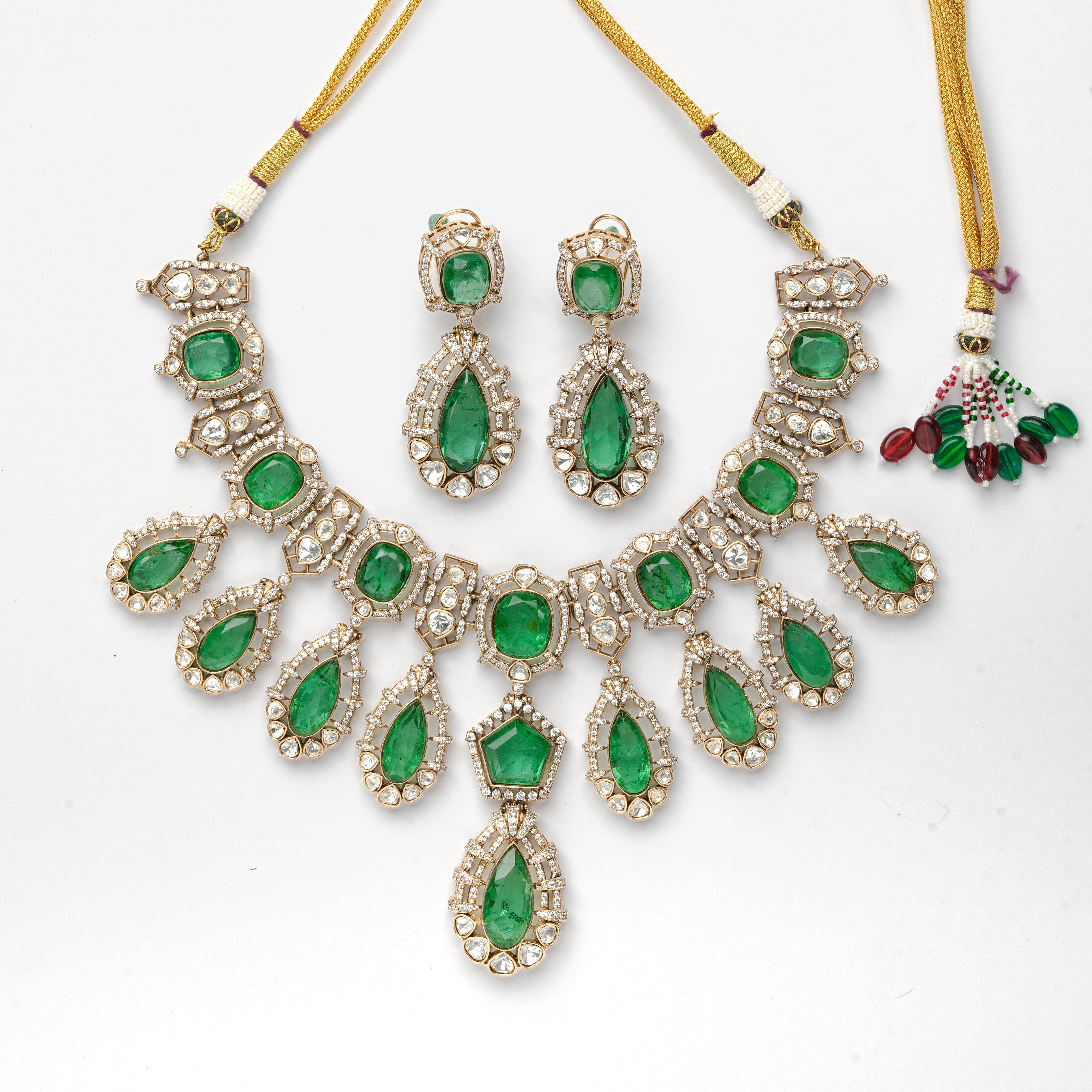 A traditional Indian jewelry set featuring a statement necklace, matching earrings, and a small hanging charm. The set is made of gold-plated metal with vibrant green gemstone accents and clear rhinestone borders, displayed on a clean white background.