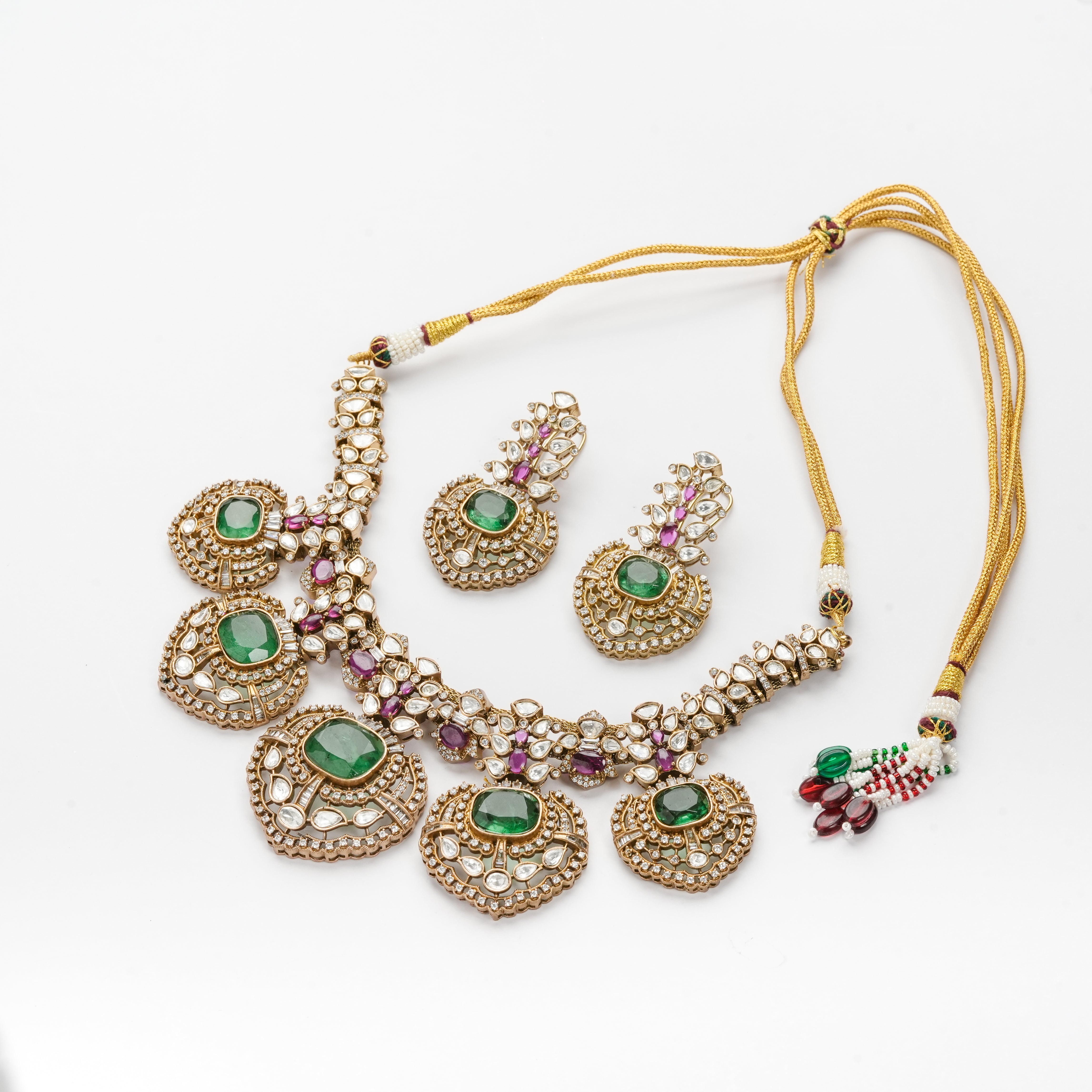 An ornate gold-toned traditional necklace set with matching earrings, featuring green stones and small pink gemstone accents on a white background.