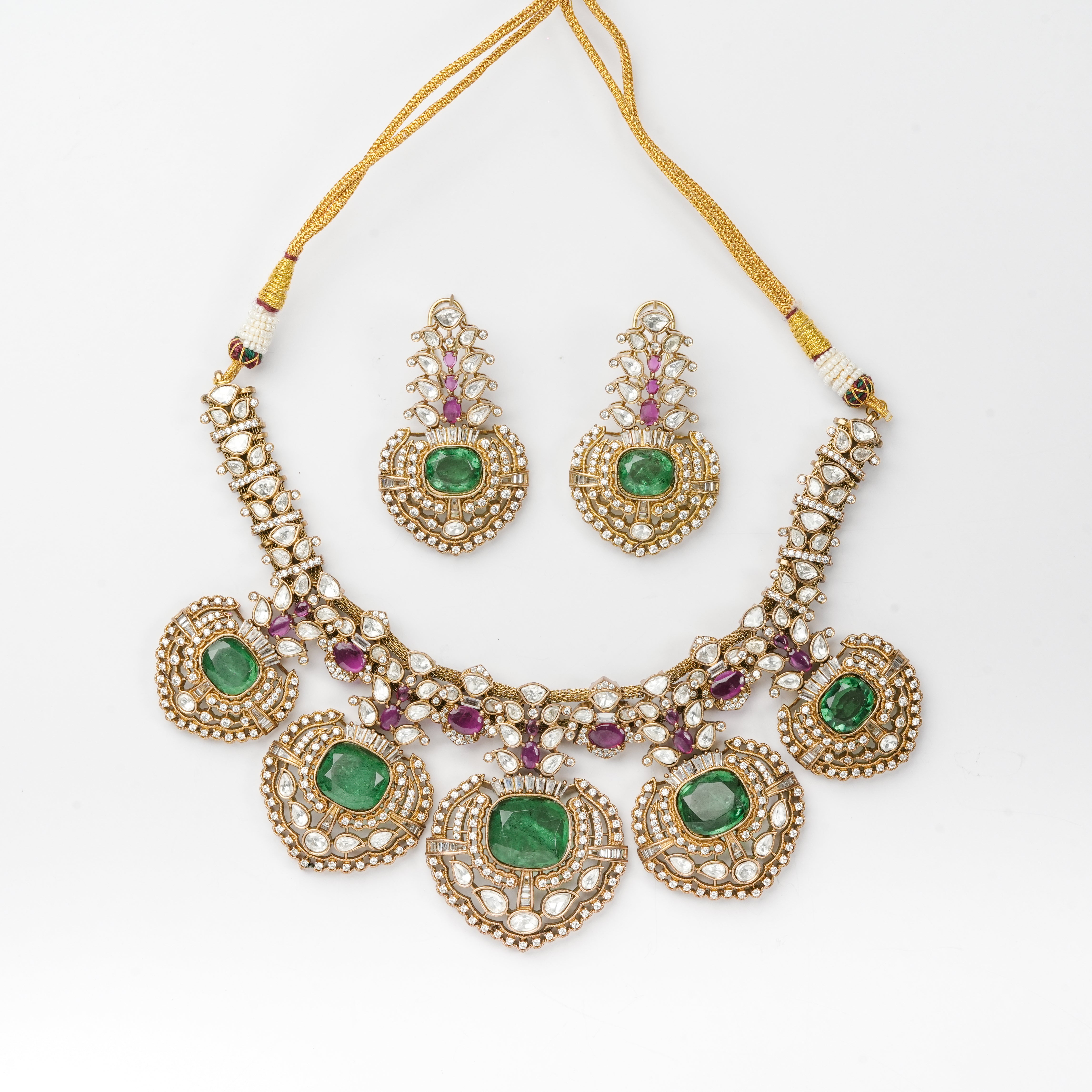 An ornate gold-toned traditional necklace set with matching earrings, featuring green stones and small purple accent stones, displayed on a clean white background.