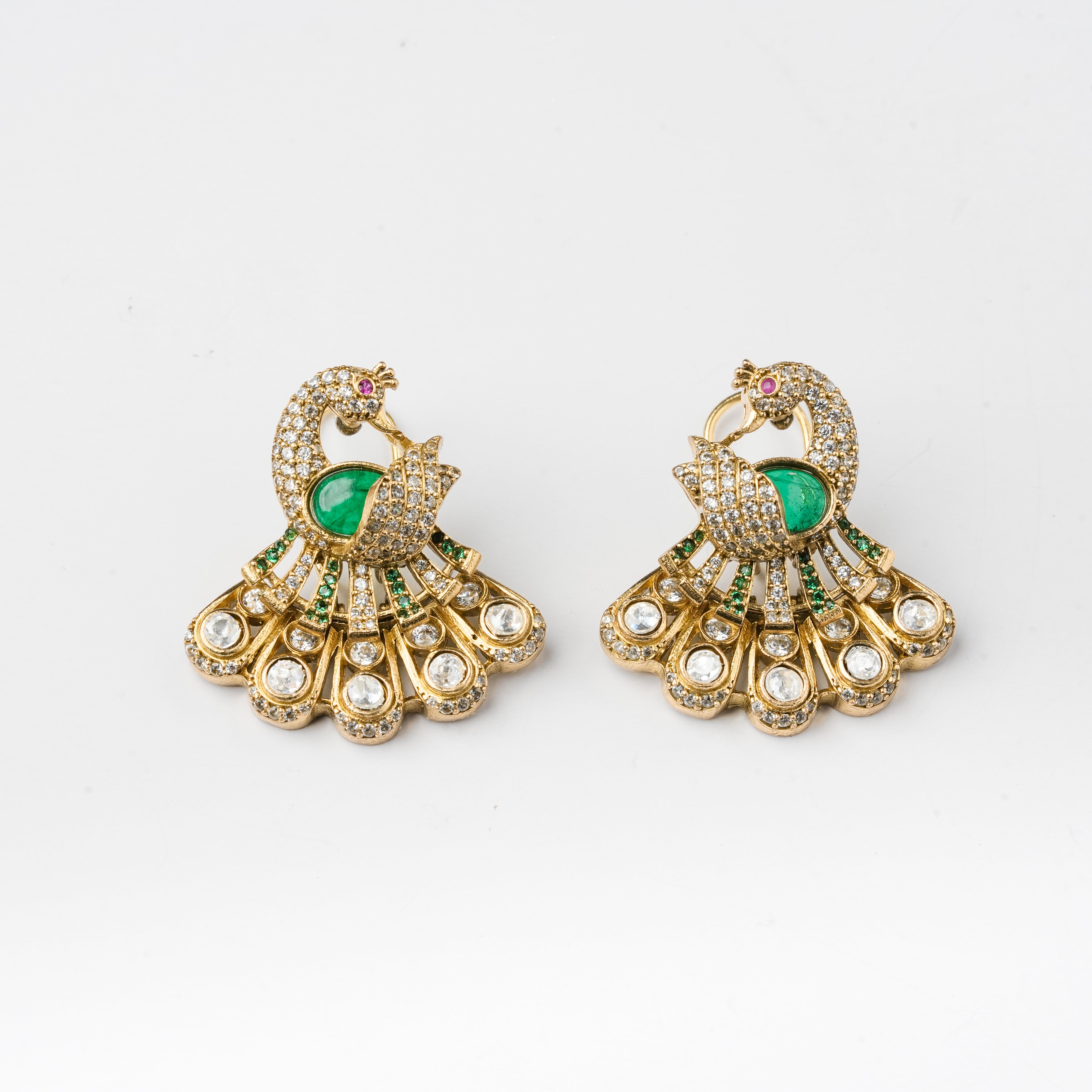 A pair of gold-toned peacock jhumka earrings embellished with green emerald-cut stones, pavé-set clear stones, and small pink accent stones, displayed on a clean white background.