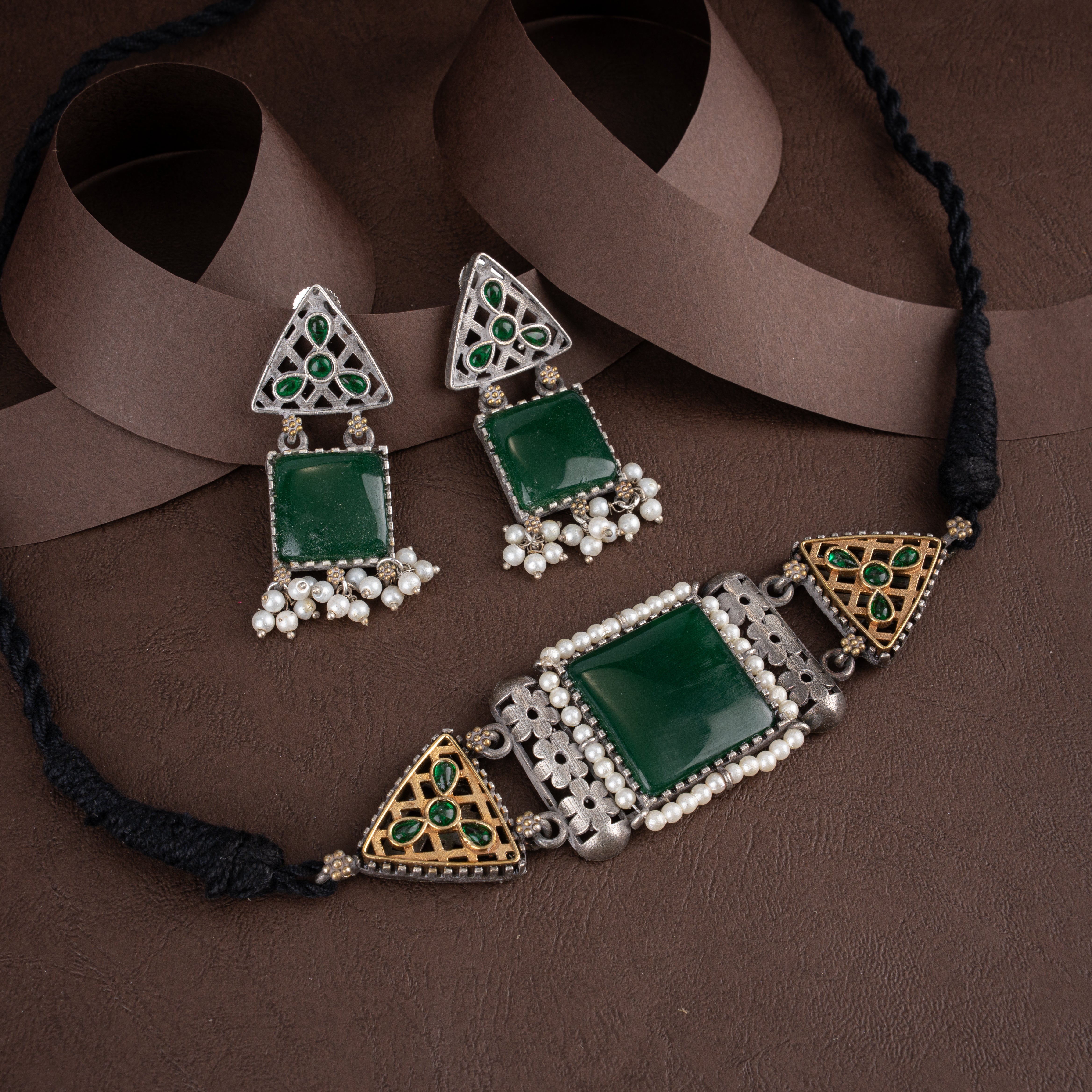 Dual-tone oxidised silver and antique gold choker necklace set featuring emerald green center stones, pearl beads, and matching drop earrings on a brown background.