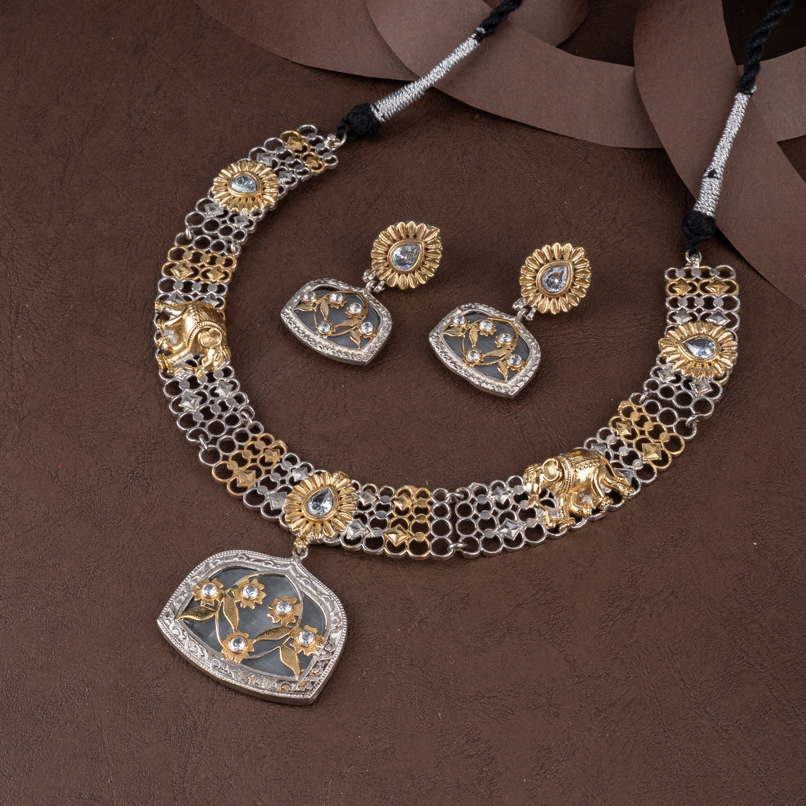 Dual-tone oxidised silver and gold mesh collar necklace set featuring elephant motifs, floral stones, and matching drop earrings on a brown background.