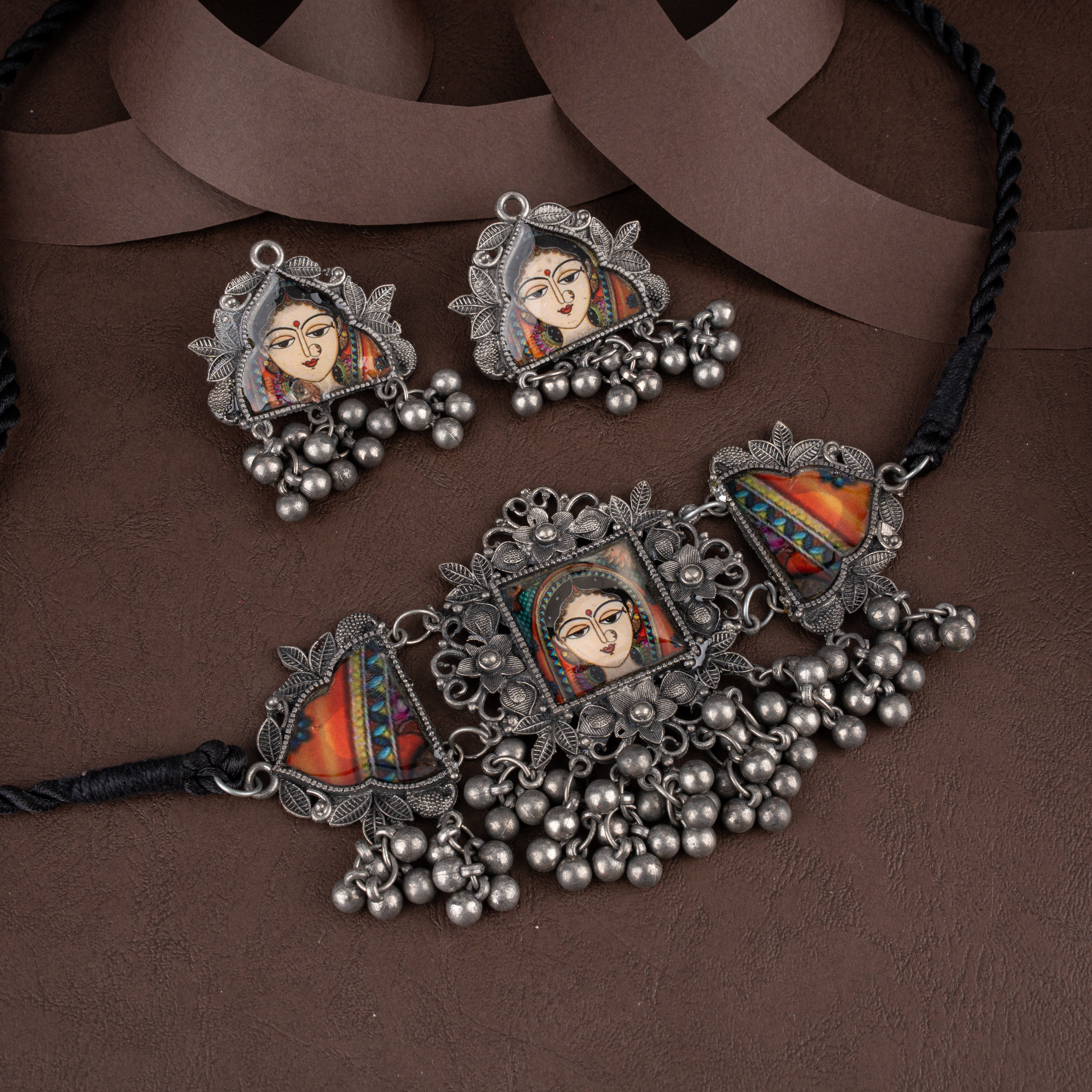 Oxidised silver choker necklace and matching earrings featuring hand-painted traditional bride portraits and hanging ghungroo beads on a brown background.
