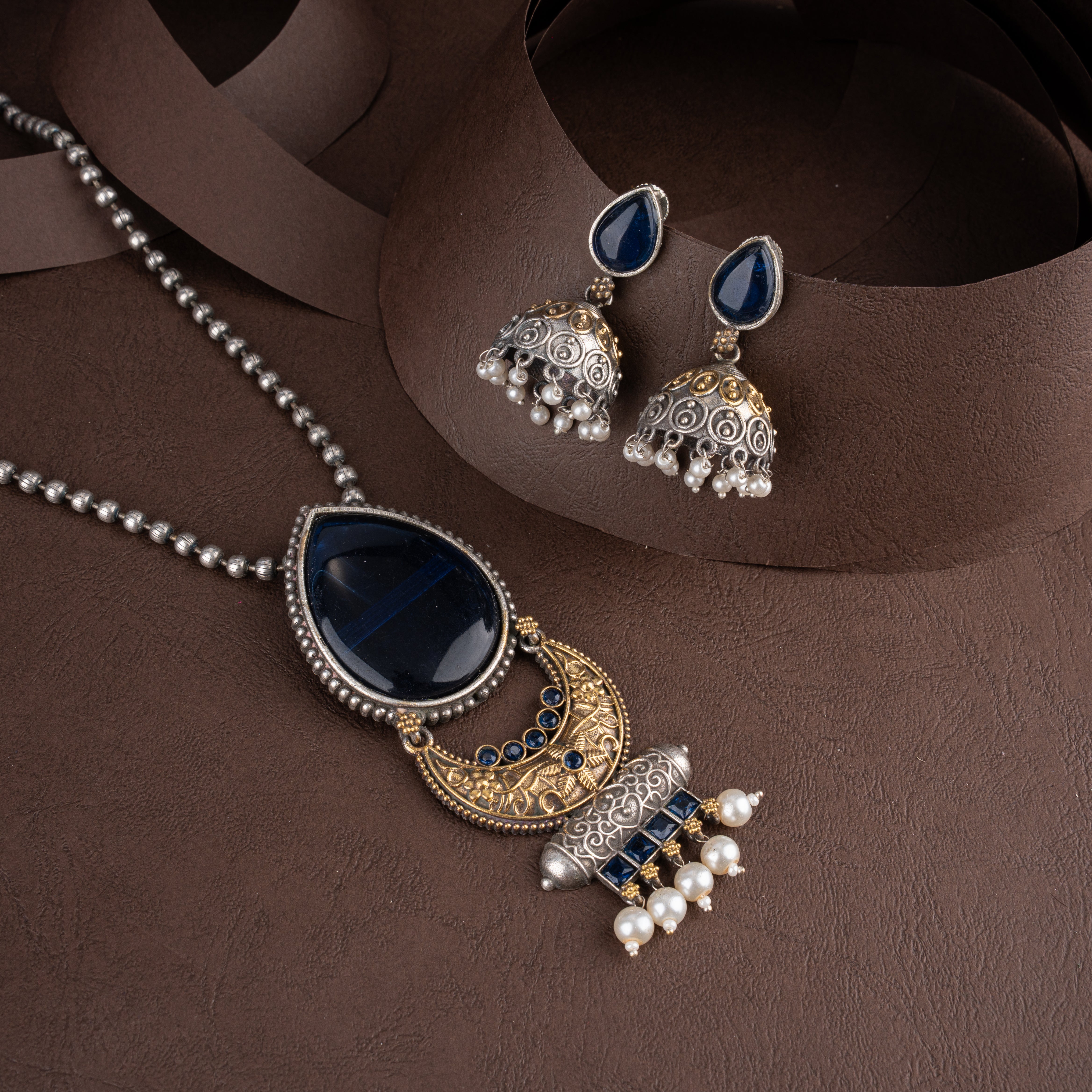 Dual-tone oxidised silver and gold pendant necklace set featuring a teardrop navy blue stone, peacock motifs, pearl drops, and matching jhumkas on a brown background.