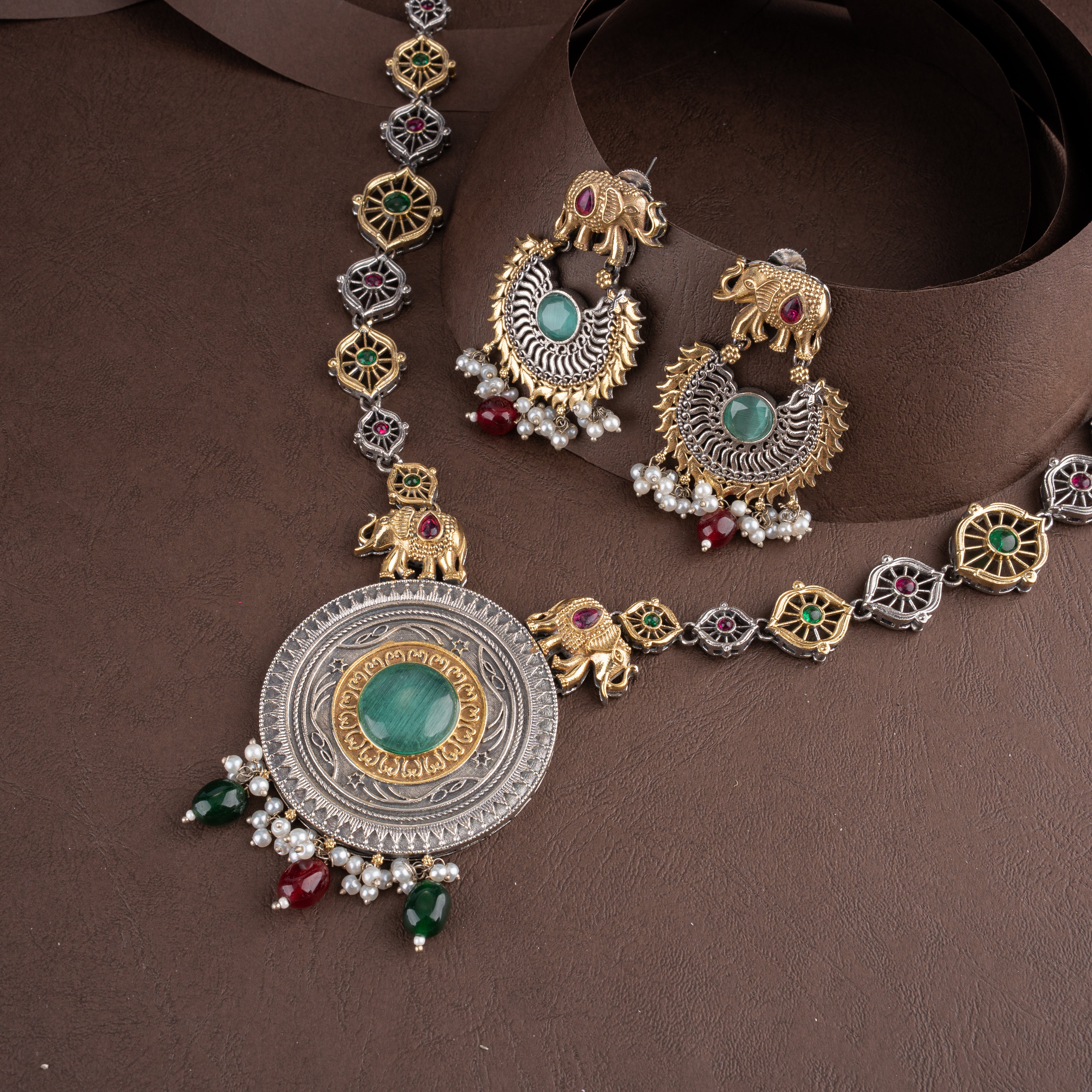 Dual-tone oxidised silver and gold long necklace set featuring royal elephant motifs, green center stones, and matching drop earrings on a brown background.