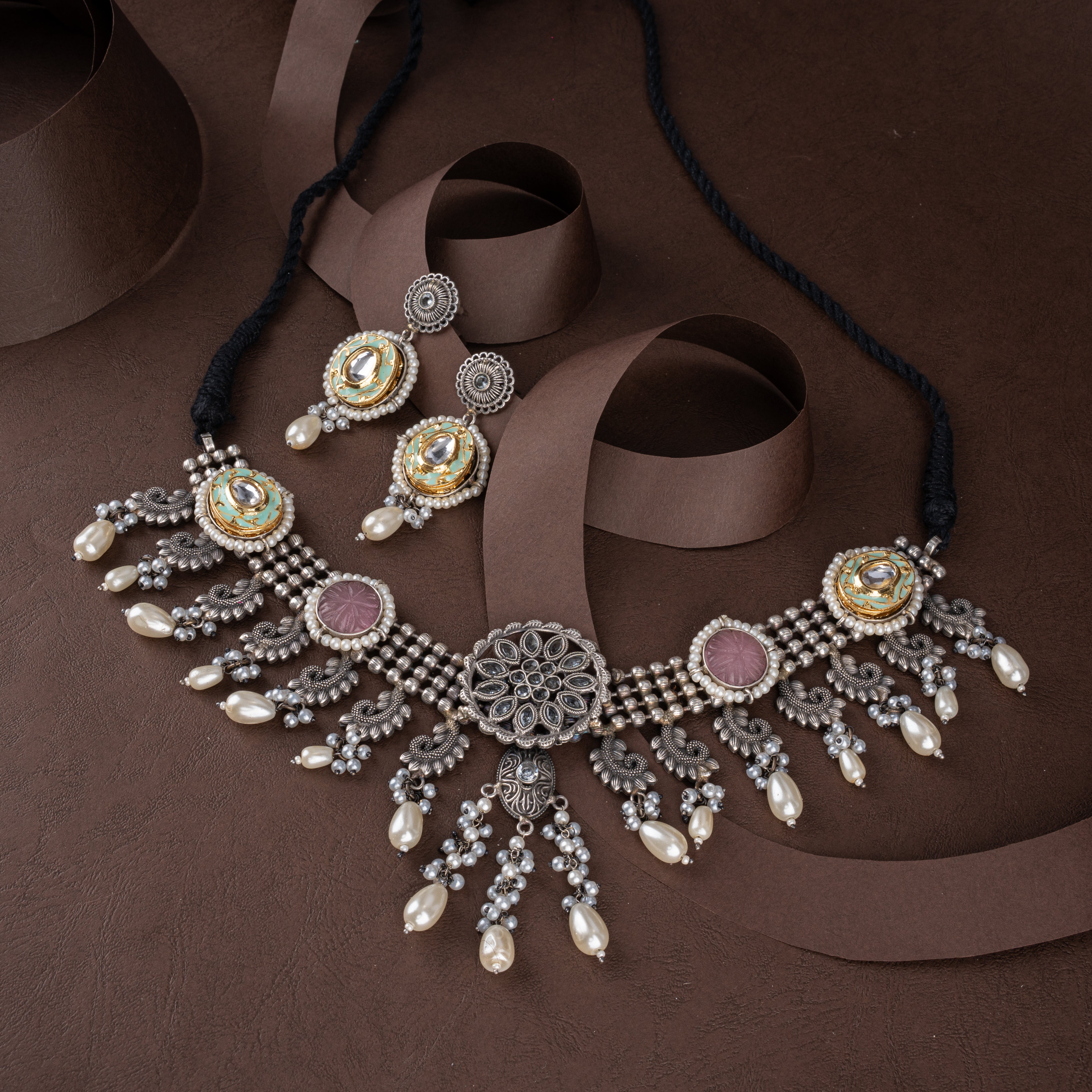Dual-tone oxidised silver choker necklace set featuring meenakari kundan medallions, floral carvings, pearl drops, and matching earrings on a brown background.