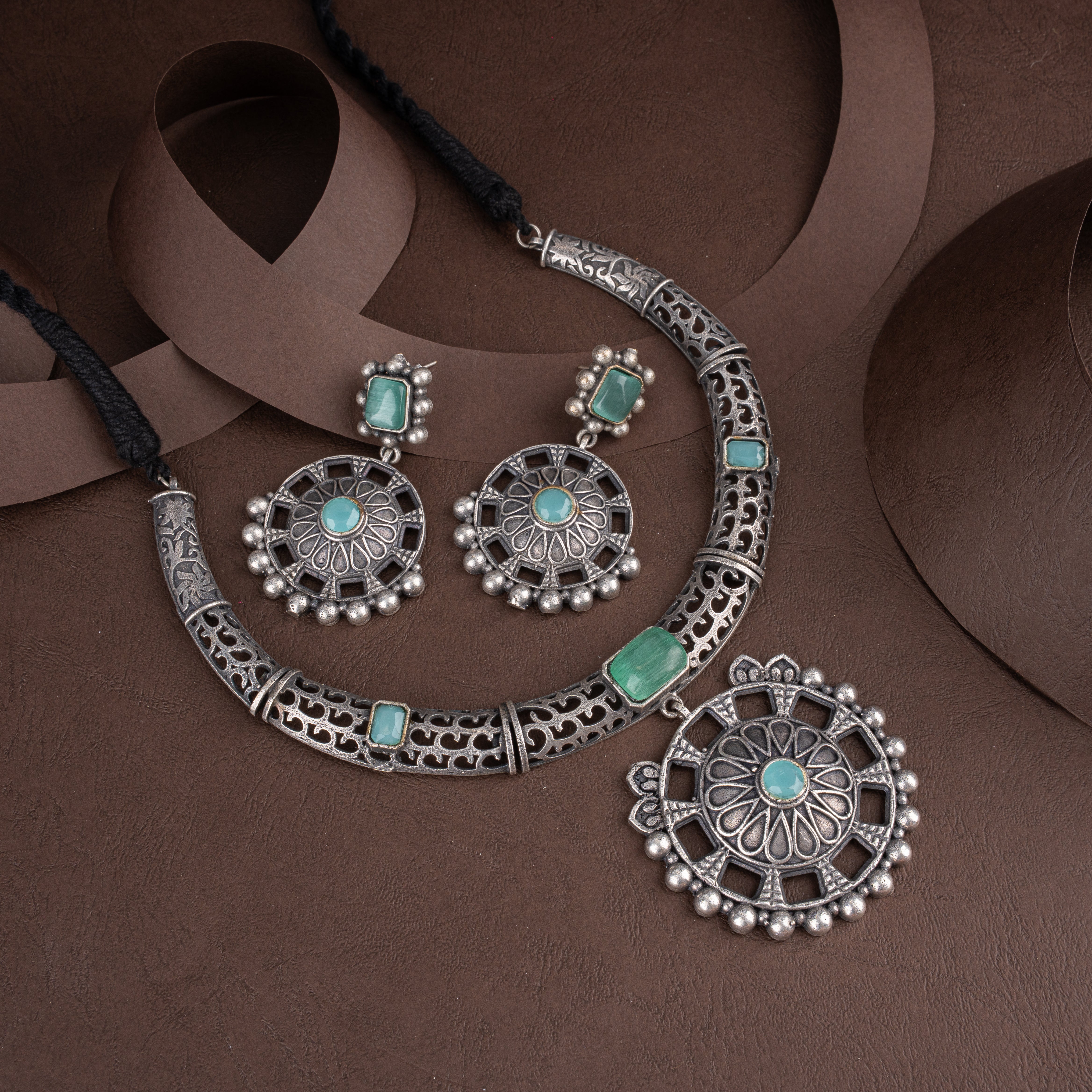 Oxidised silver hasli necklace set studded with mint green stones, featuring a round filigree pendant and matching drop earrings on a brown background.