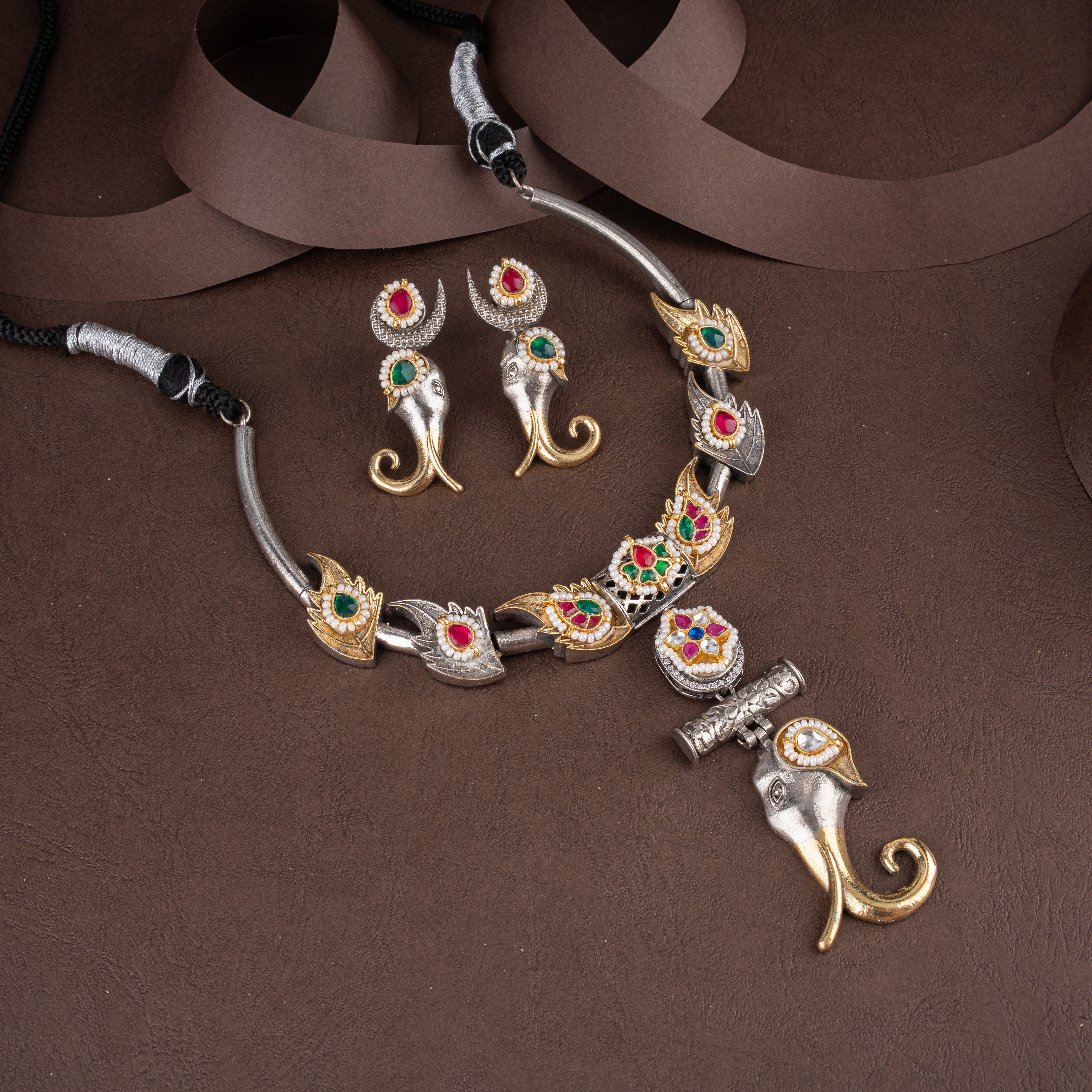 Dual-tone oxidised silver and antique gold hasli necklace set featuring an elephant trunk pendant, pearl borders, and matching drop earrings on a brown background.