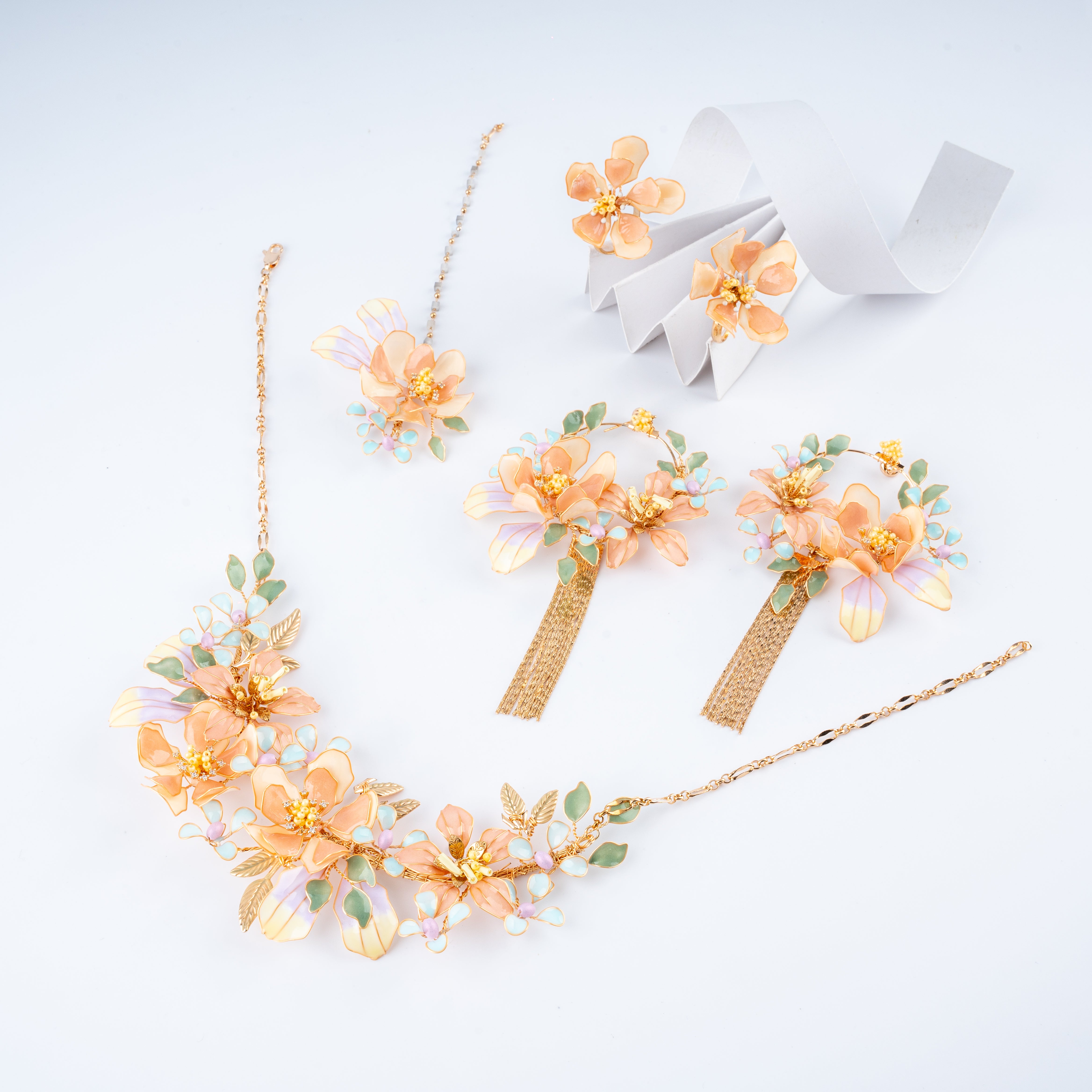 A floral jewelry set on a clean white background, including a necklace, matching earrings, and a folded white ribbon. The jewelry features peach and yellow flower petals with green leaves and iridescent pastel accents on a gold-tone chain.