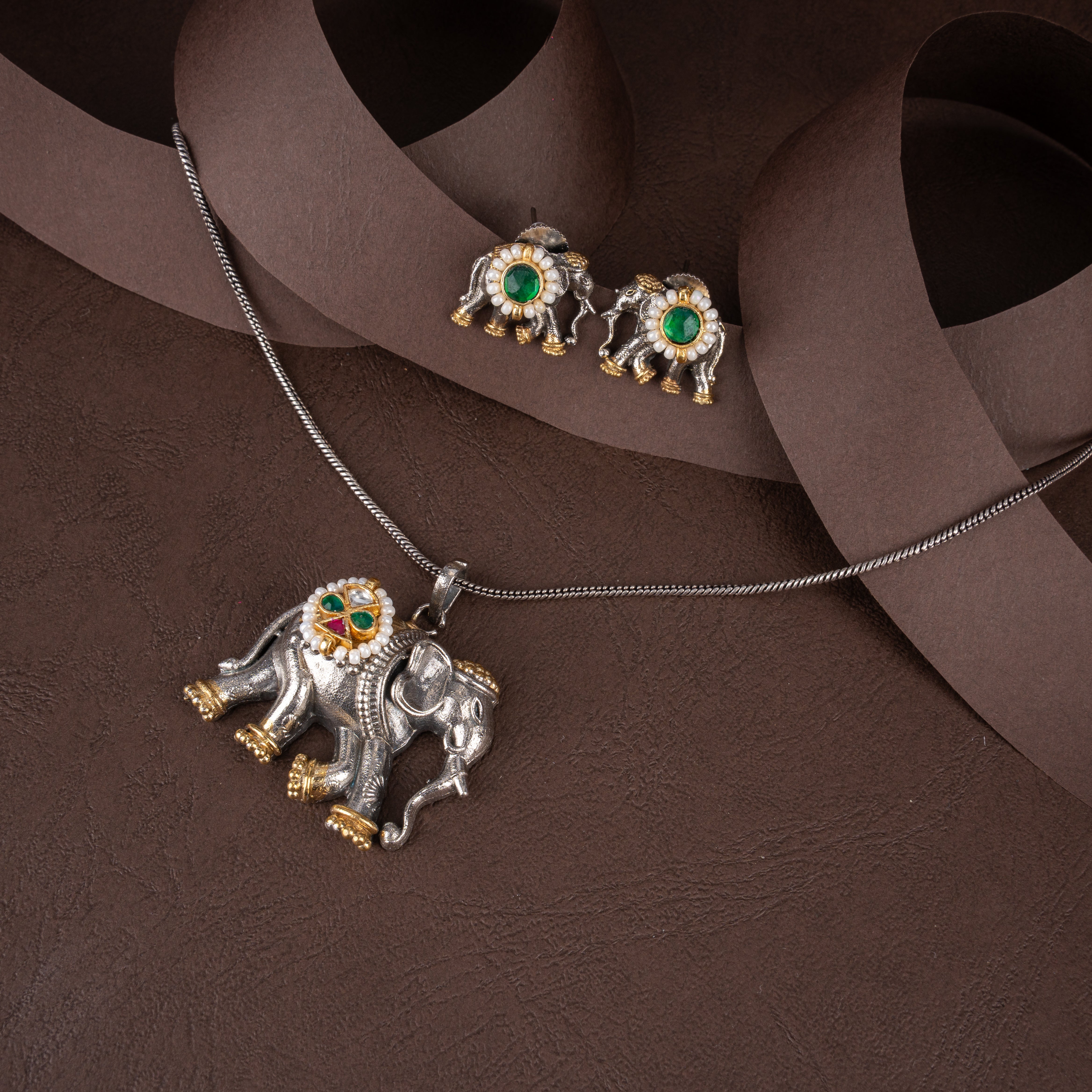 Dual-tone oxidised silver and gold elephant pendant necklace with micro pearl detailing and matching elephant stud earrings on a brown background.
