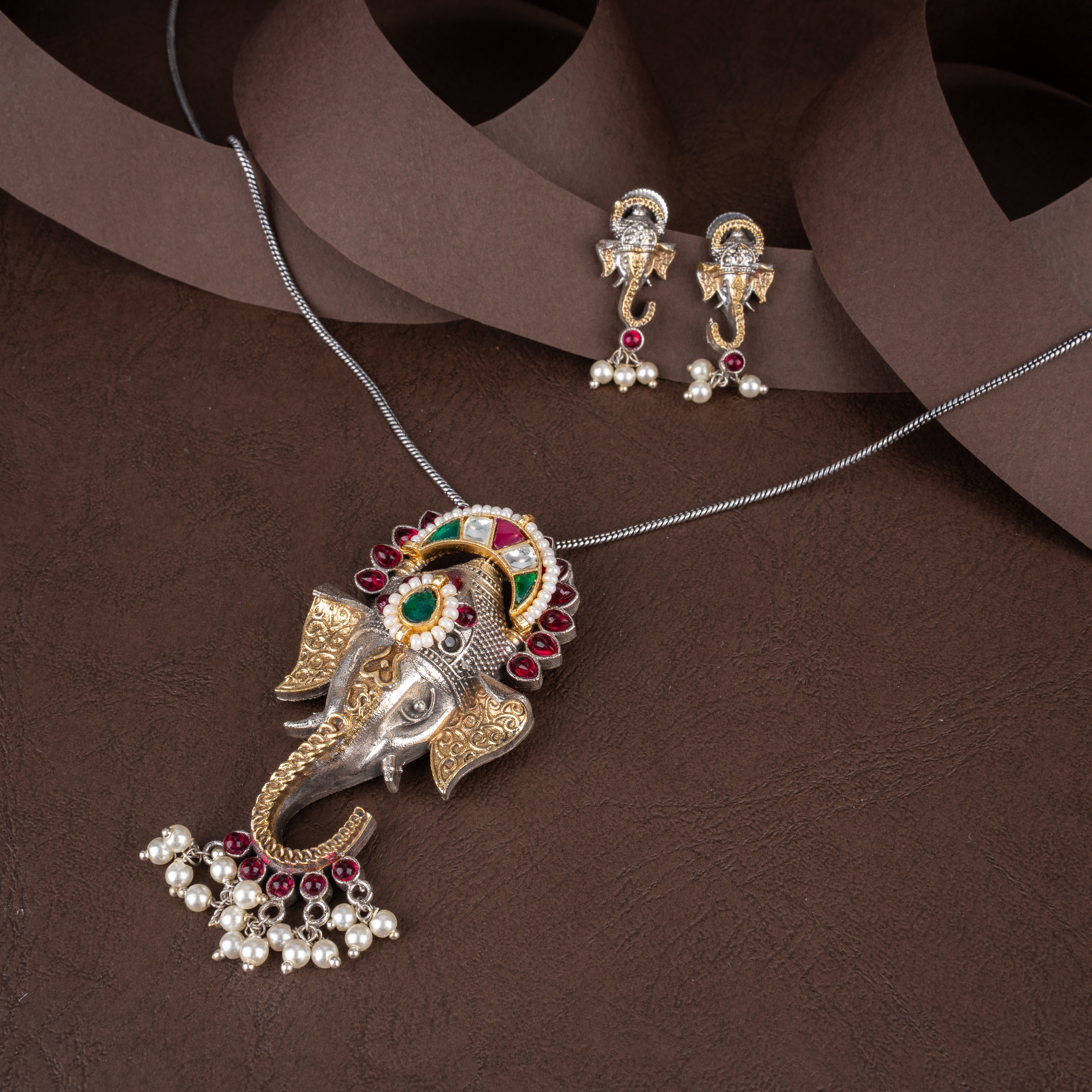 Dual-tone oxidised silver and gold Lord Ganesha pendant necklace with pearl clusters and matching Ganesha drop earrings on a brown background.