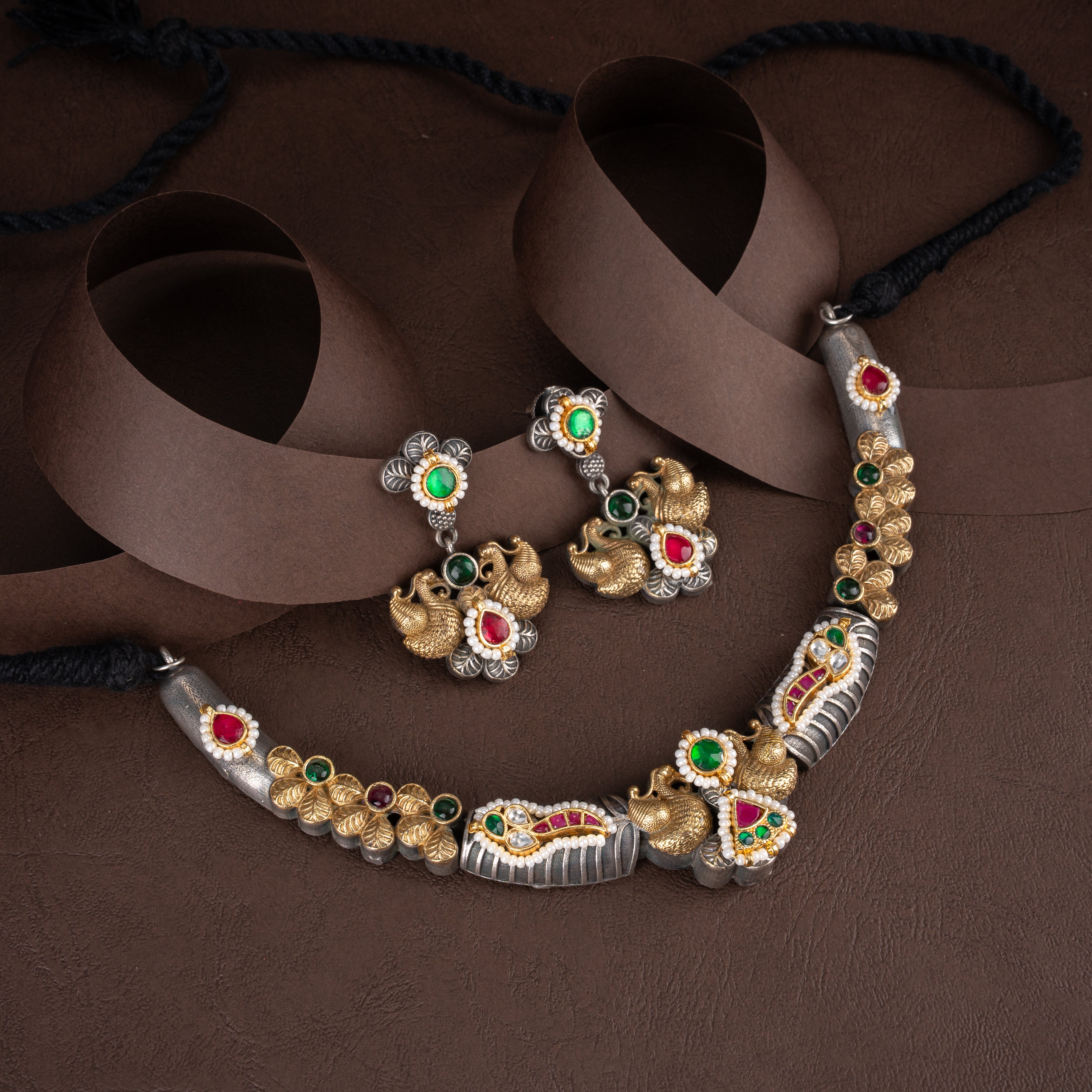 Dual-tone oxidised silver and gold peacock hasli necklace set with floral leaf accents, gemstone inlays, and matching earrings on a brown background.