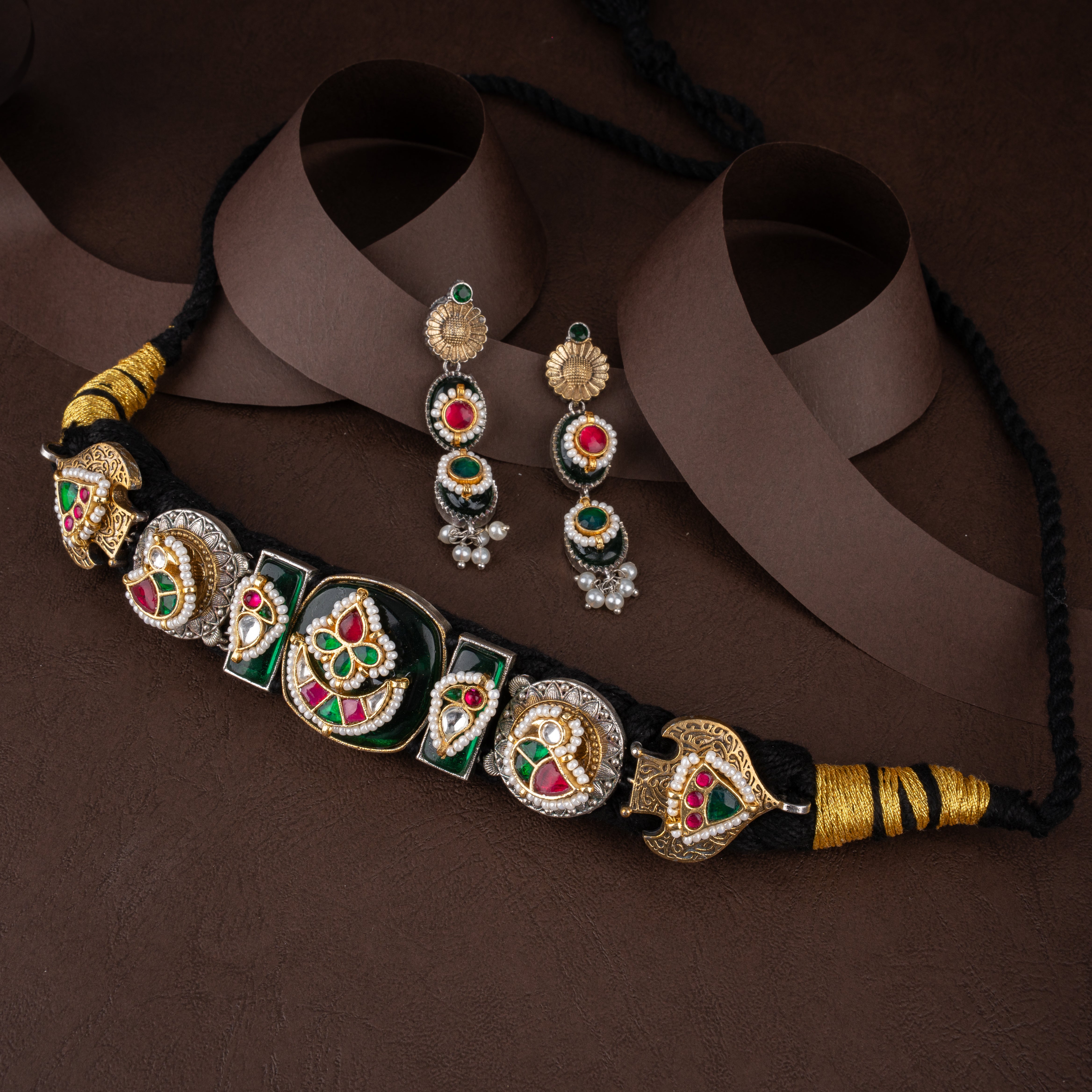 Dual-tone oxidised silver and gold tribal fabric choker necklace set with gemstone motifs, pearl borders, and matching drop earrings on a brown background.