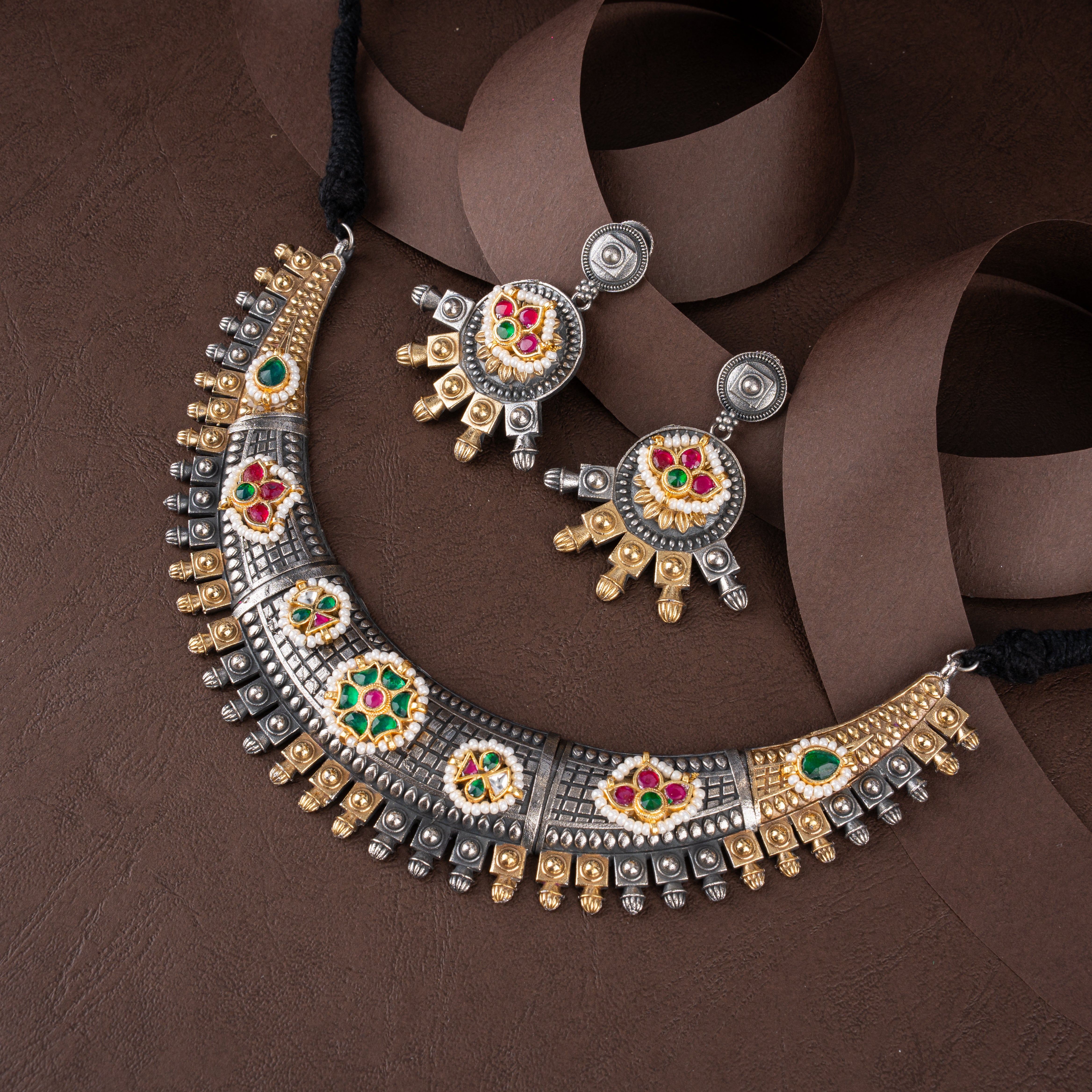 Dual-tone oxidised silver and gold collar necklace set featuring floral stone medallions, fluted edge spikes, and matching drop earrings on a brown background.