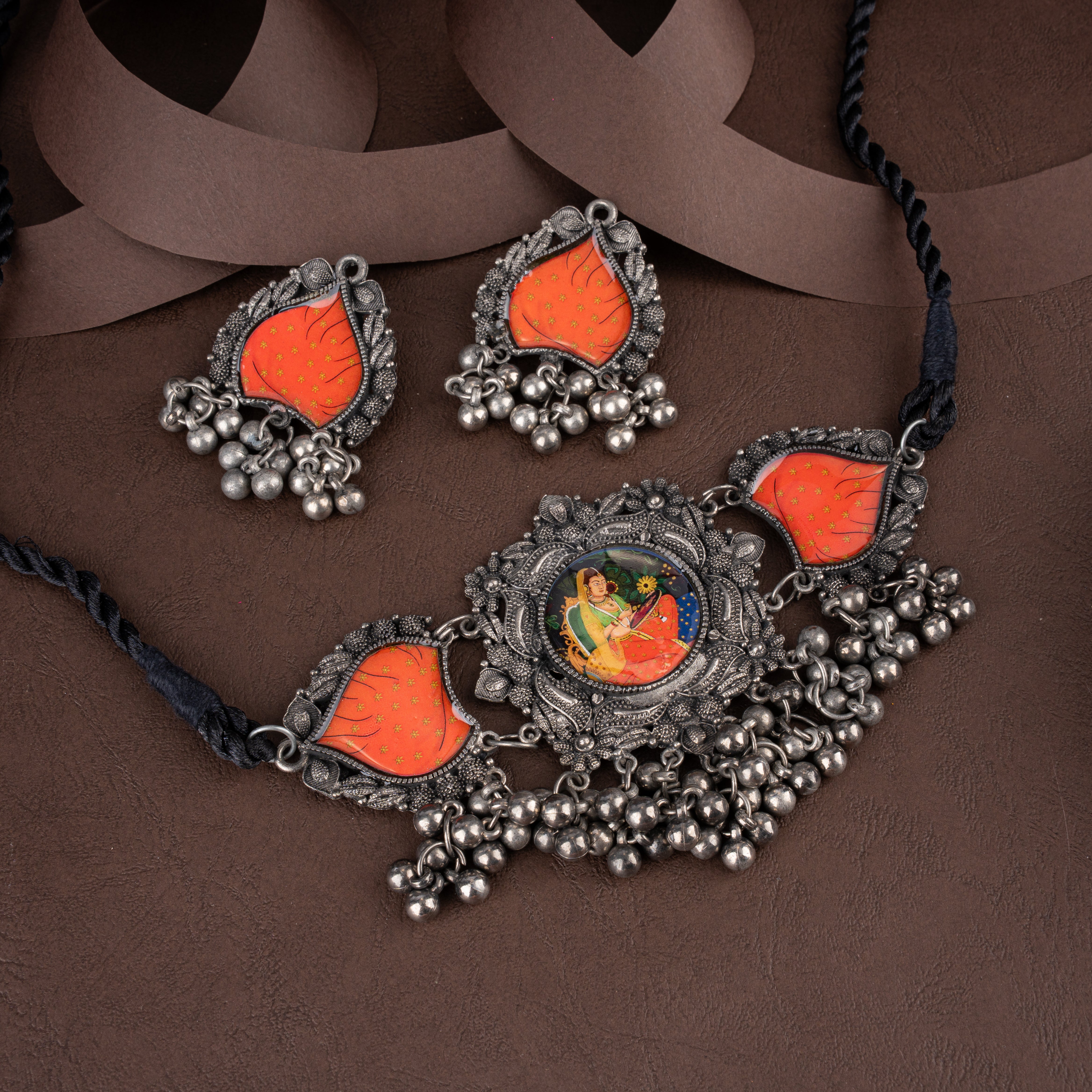 An ornate silver-toned jewelry set featuring a necklace and matching earrings with bright orange inlay panels, silver bead clusters, and a central painted motif of a seated figure, displayed on a brown textured surface with draped brown ribbon.