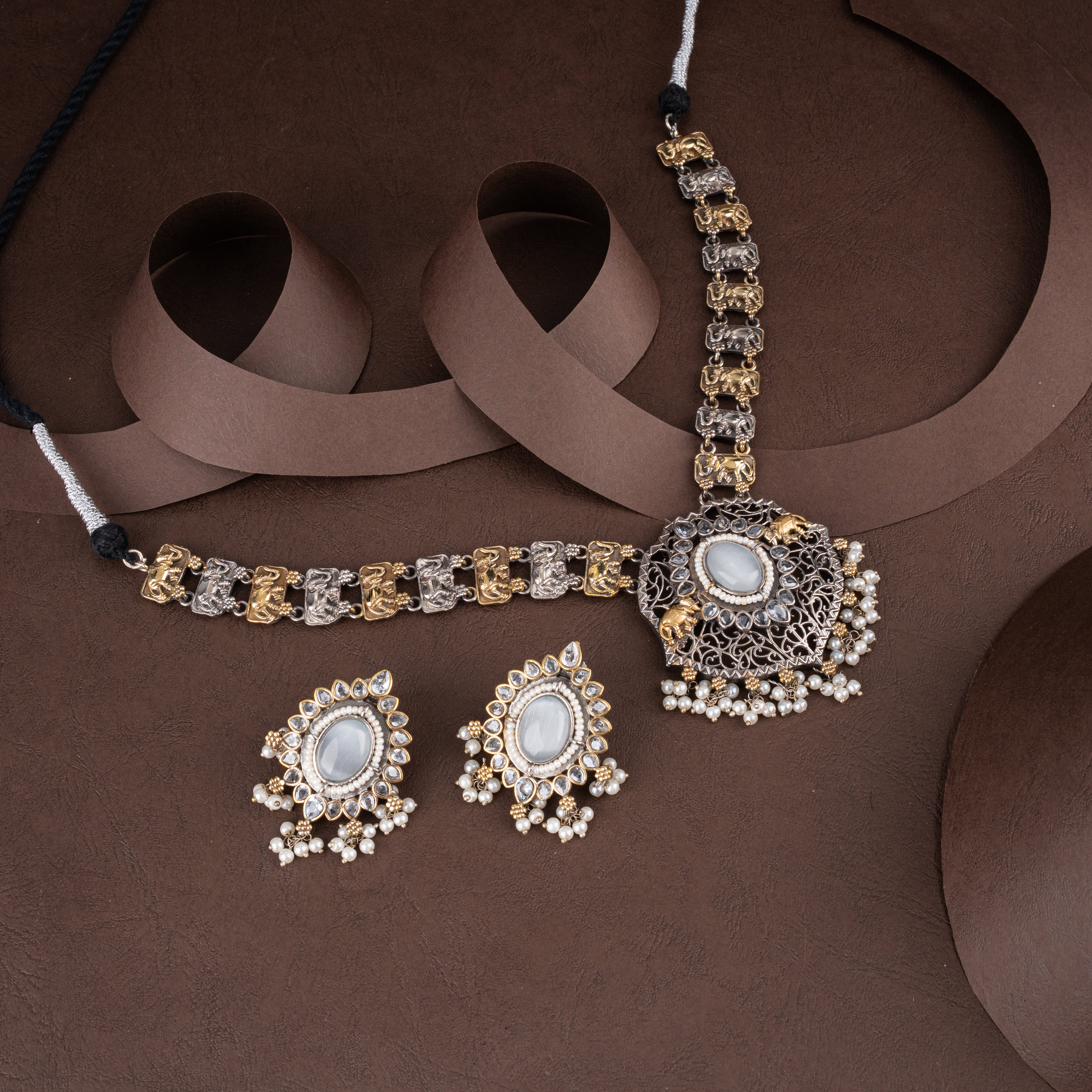 Dual-tone oxidised silver and gold elephant motif choker necklace set featuring white center stones, pearl drop tassels, and matching earrings on a brown background.