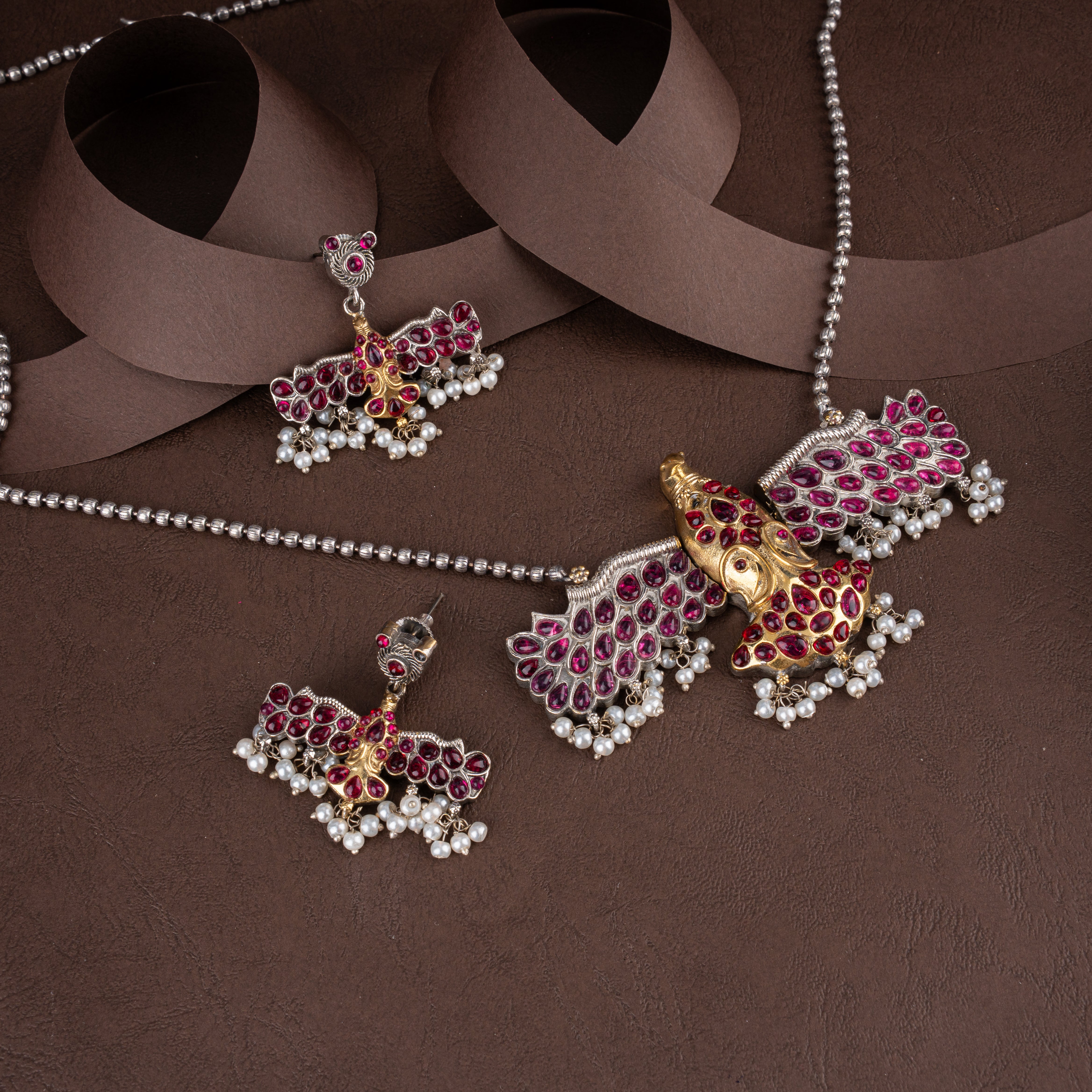 Dual-tone oxidised silver and gold Lord Ganesha pendant necklace set with ruby-pink stones, seed pearl drops, and matching earrings on a brown background.