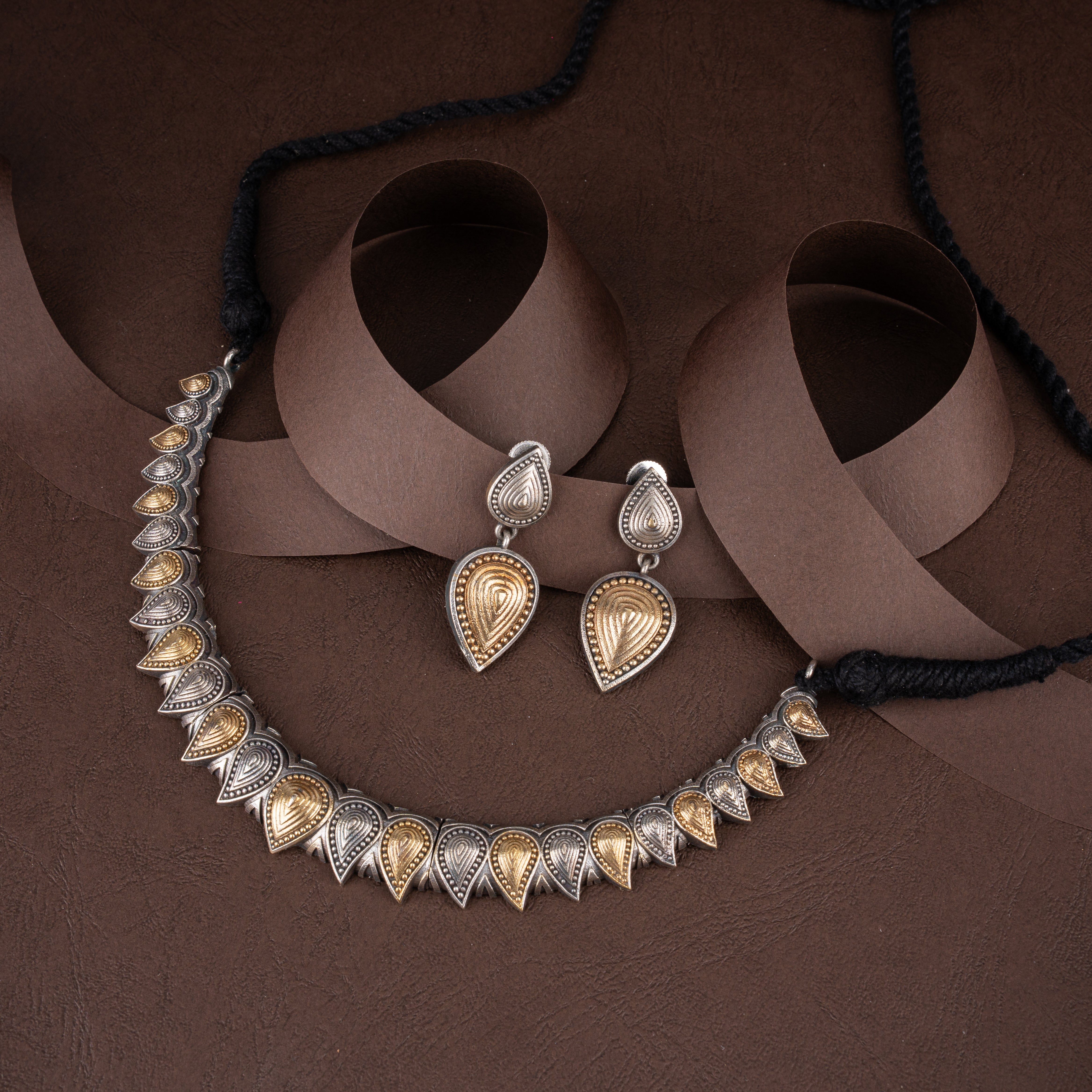 Dual-tone oxidised silver and antique gold teardrop leaf collar necklace set with matching two-tier earrings on a brown background.