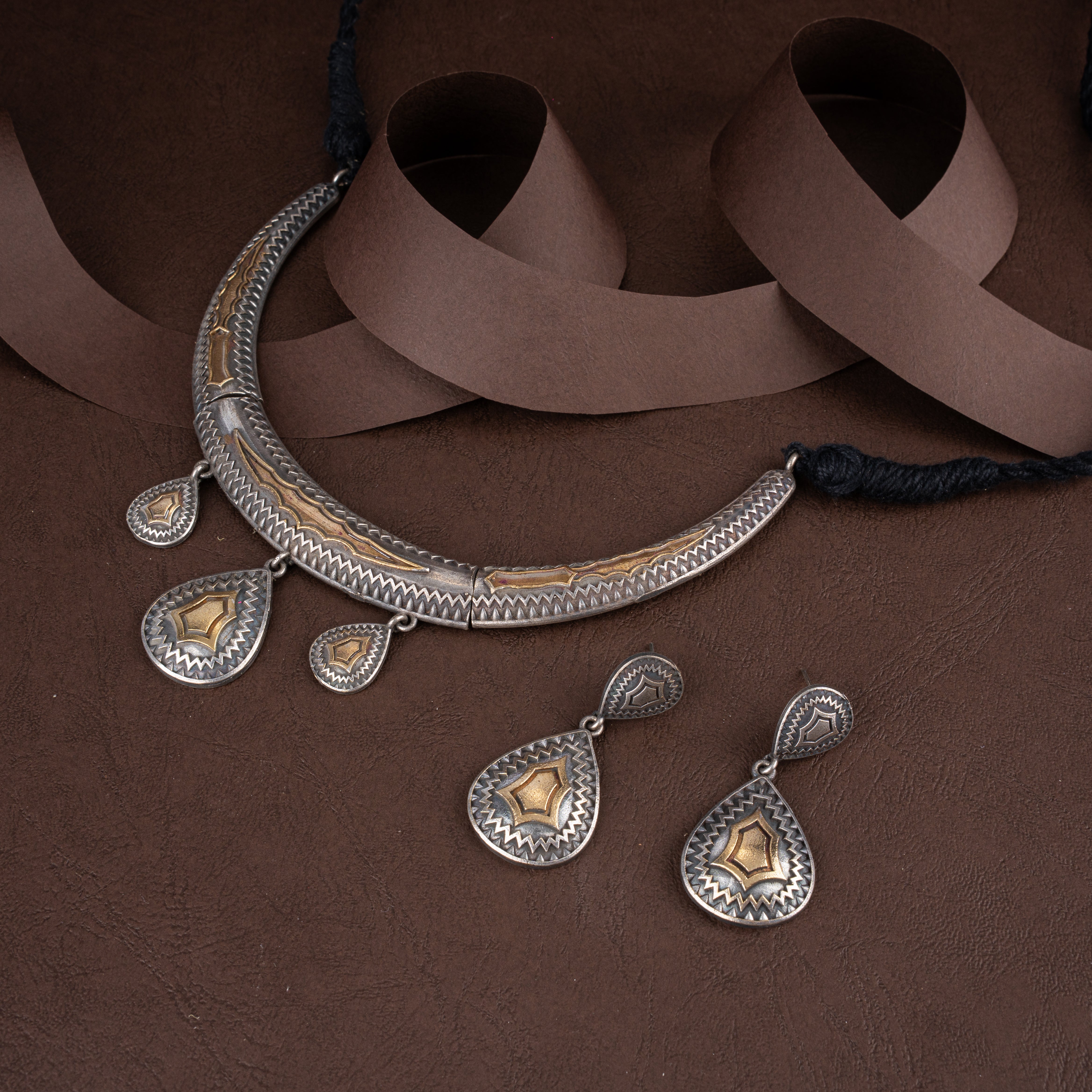 Dual-tone oxidised silver and brass tribal hasli choker necklace featuring engraved teardrop charms and matching drop earrings on a brown background.