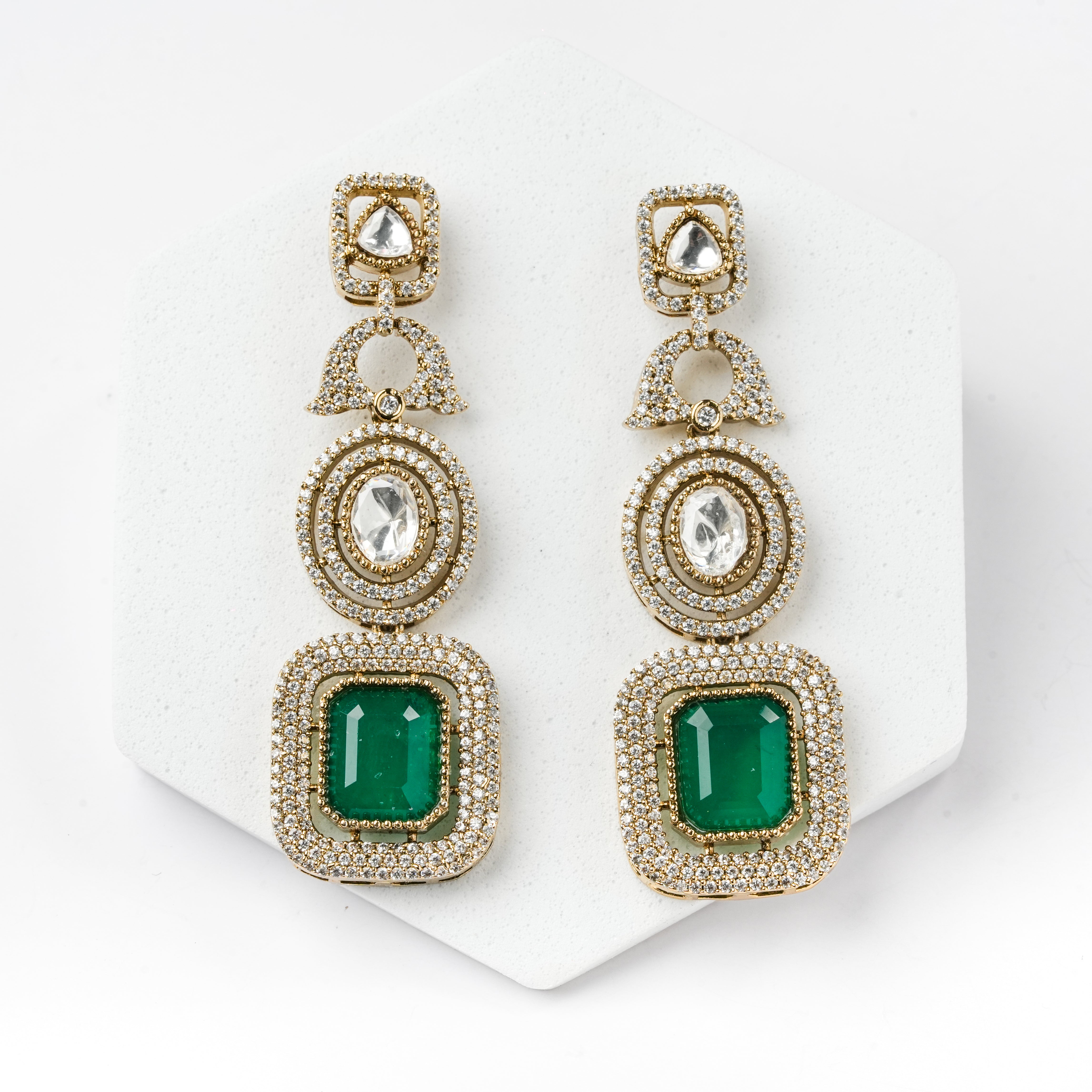 A pair of gold drop earrings with clear pavé-set stones and large emerald-cut green gemstones, displayed on a white hexagonal stand.