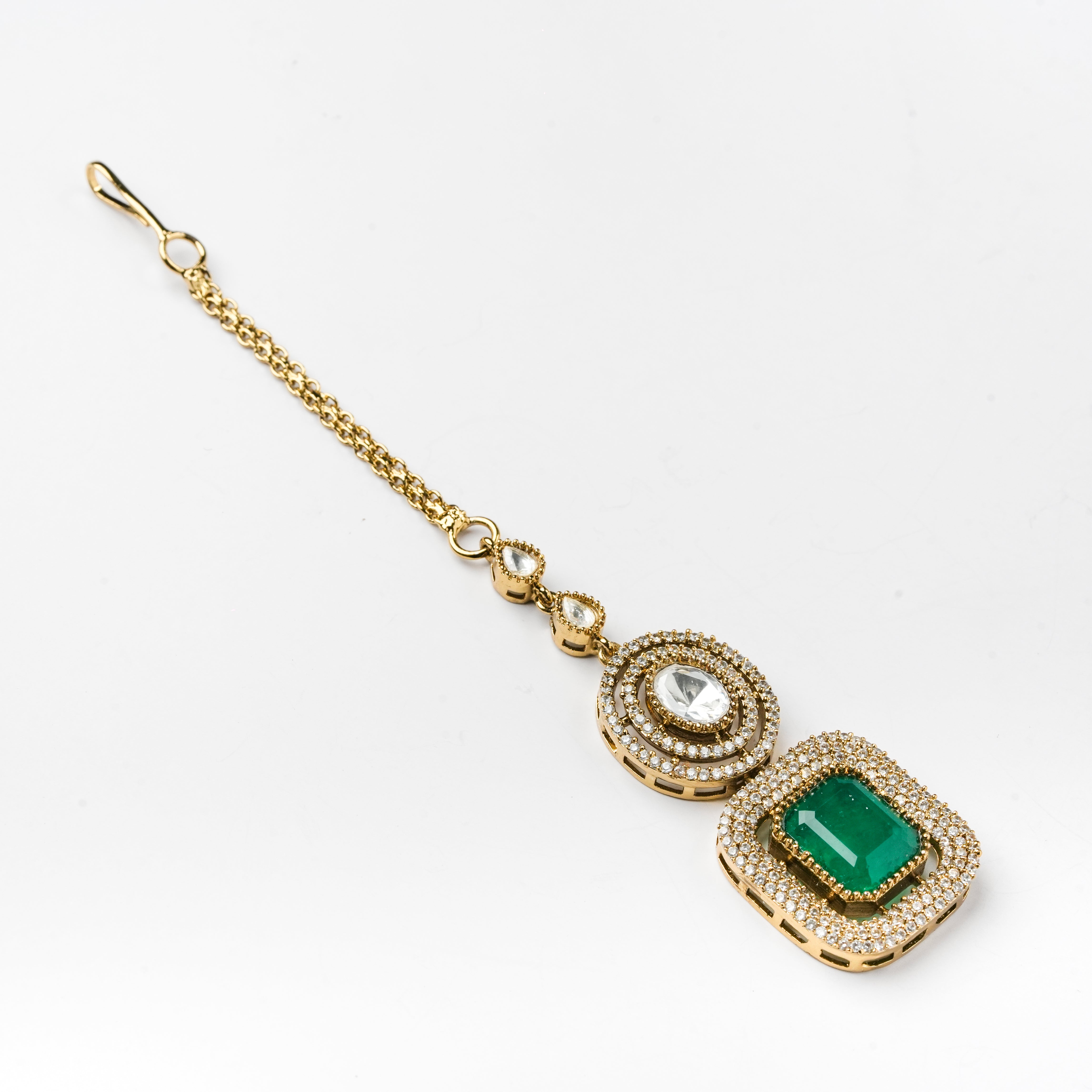 A gold-toned traditional Indian pendant necklace featuring a rectangular green emerald-cut stone surrounded by small clear stones, with a circular clear stone pendant above it, all on a delicate chain with a lobster clasp.
