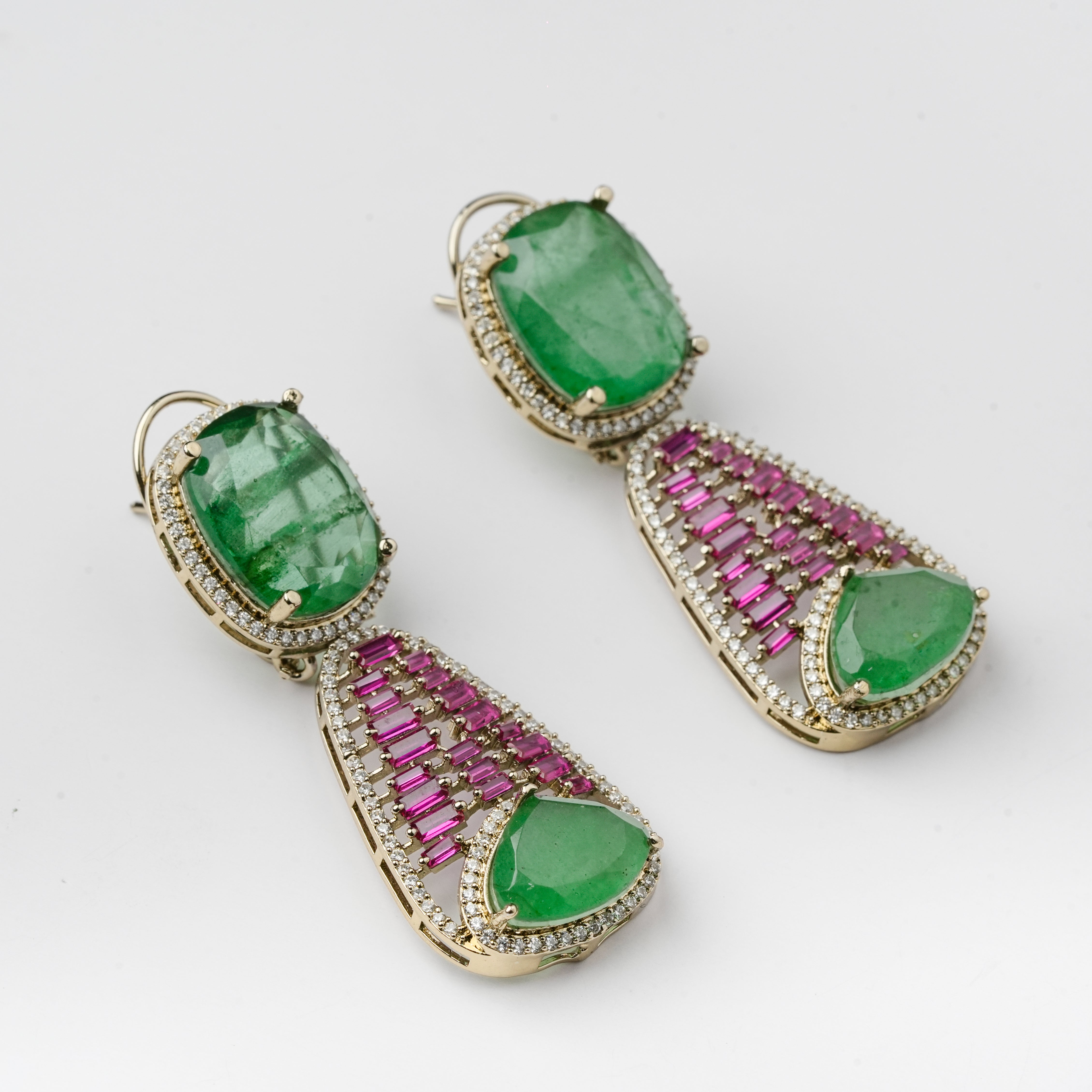 A pair of gold drop earrings featuring large green emerald-cut stones, pink baguette-cut stones, and small clear stones, shown on a light gray background.